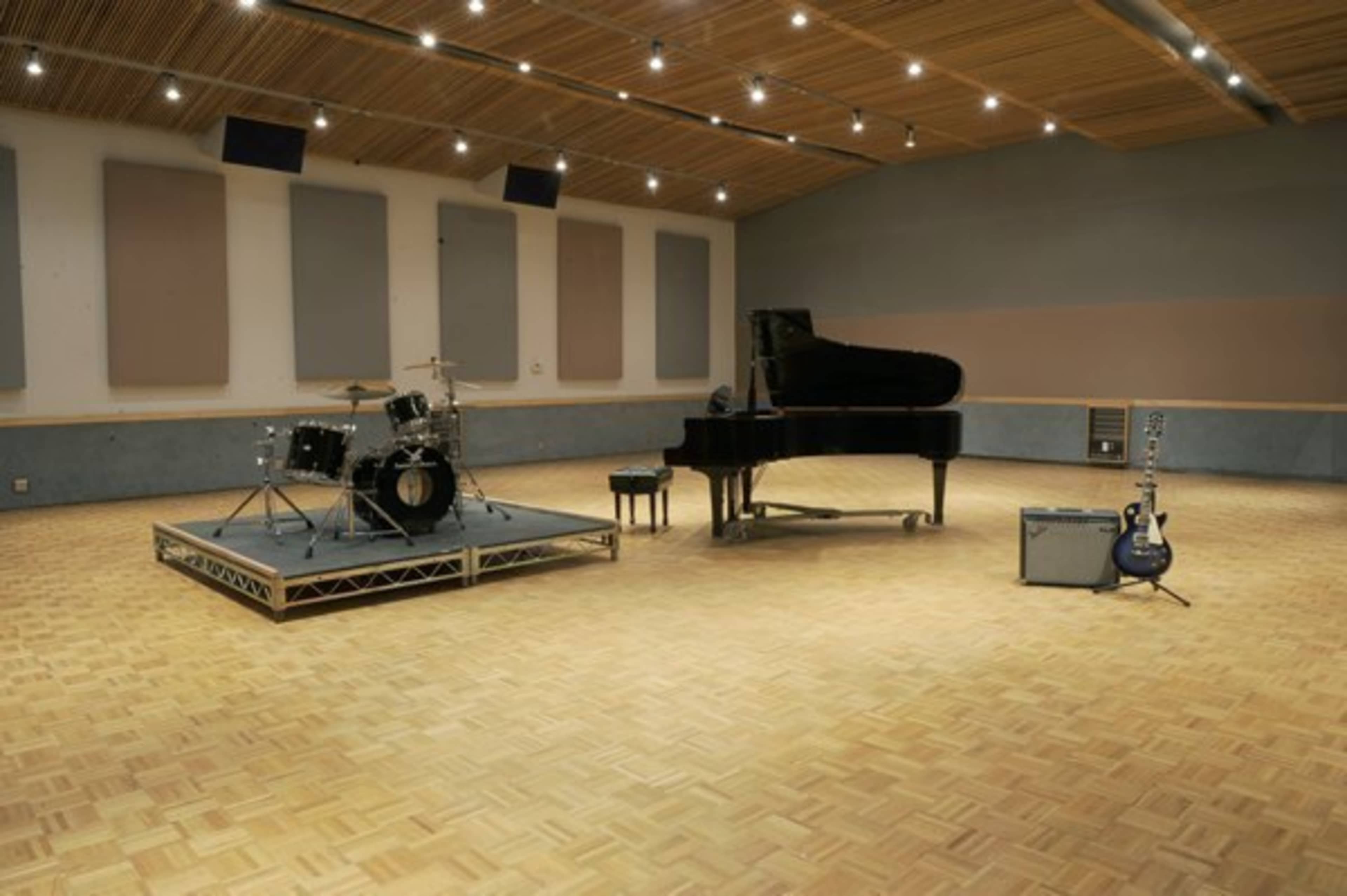 A spacious recording studio featuring a grand piano, a drum set on a raised platform, an electric guitar, and an amplifier.