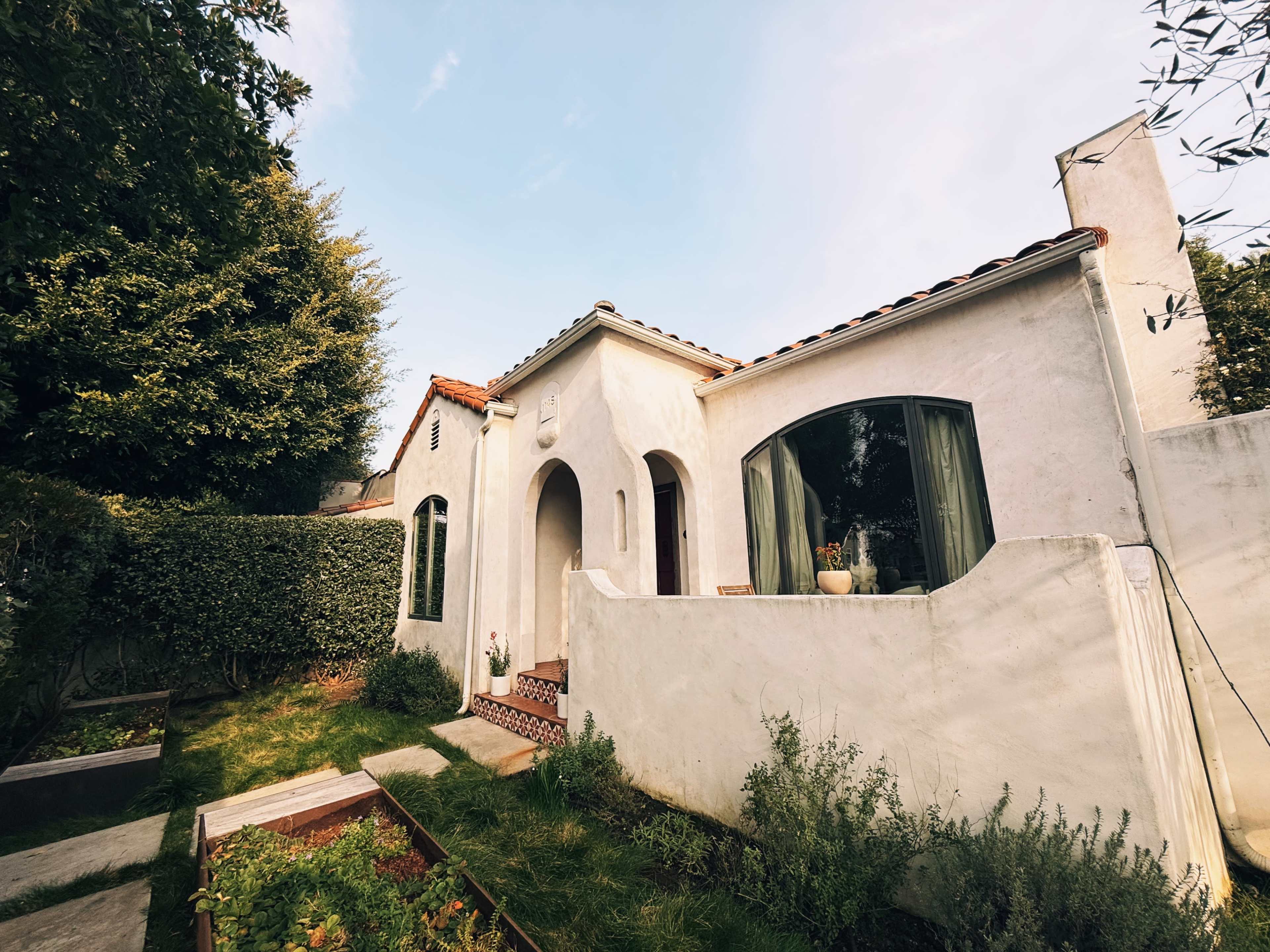 A single-story, white stucco house with a tile roof and arched windows is surrounded by greenery and a small garden.