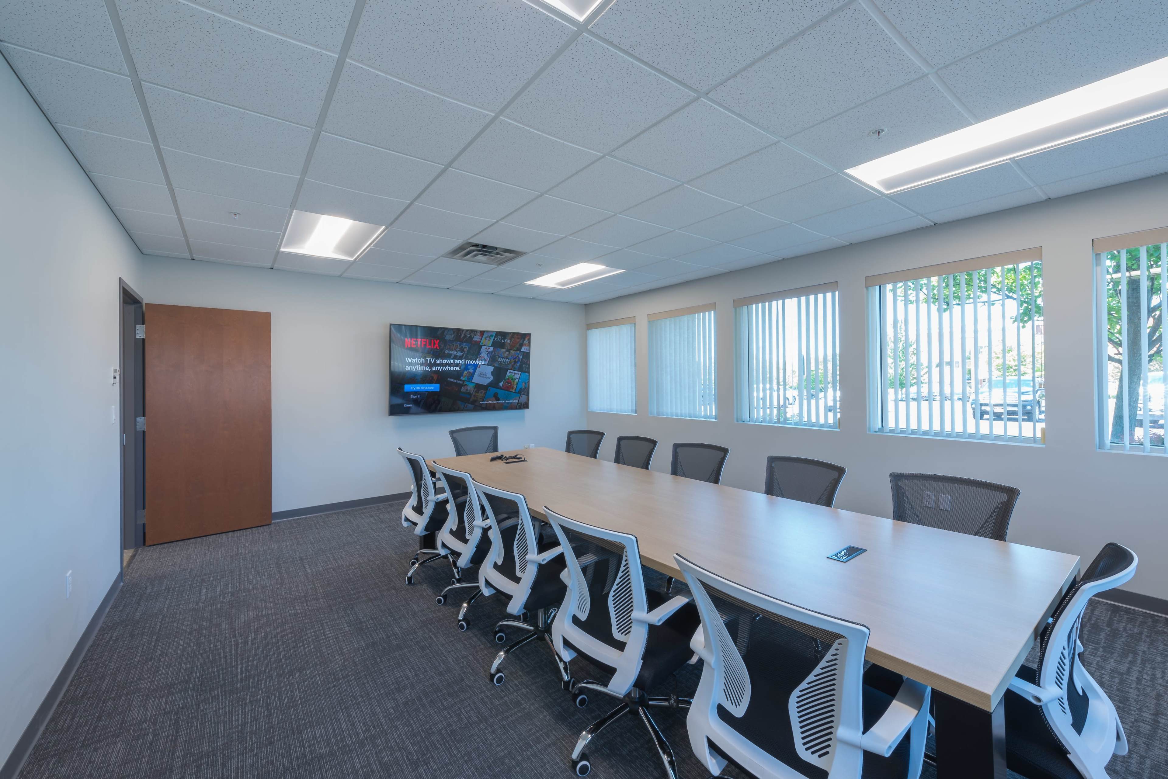 A conference room features a long wooden table surrounded by ergonomic chairs, with a large screen mounted on the wall.