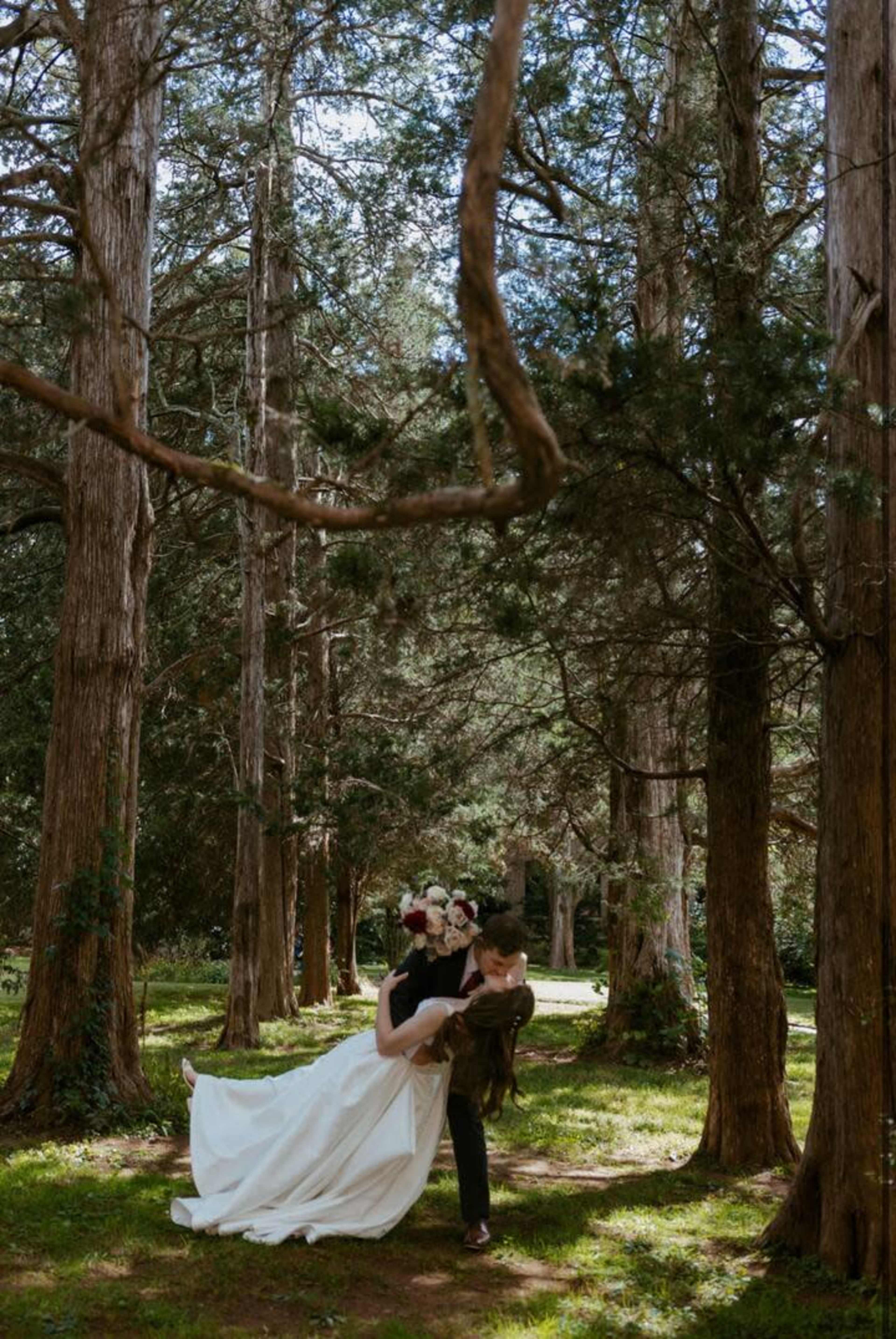 A newlywed couple poses in a forest surrounded by tall trees, with the groom lifting the bride in his arms.
