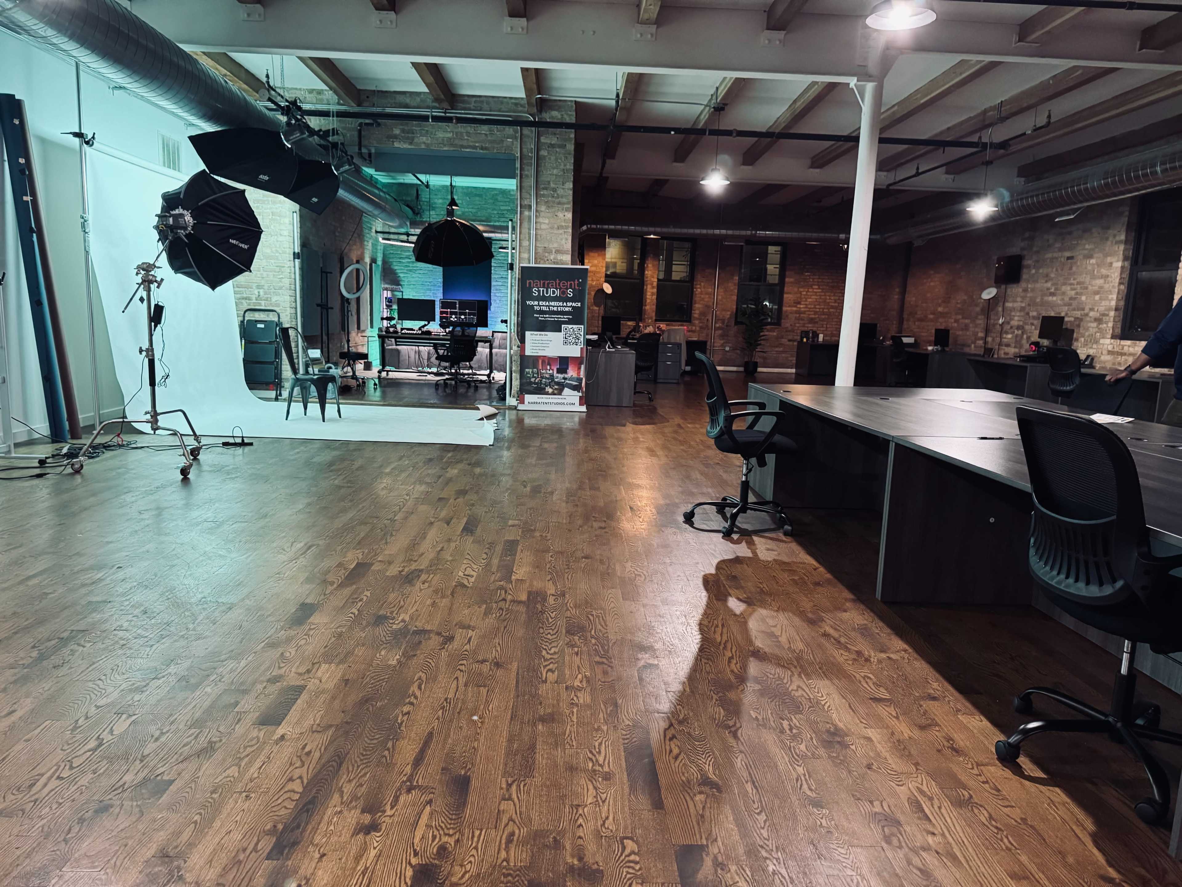 The image shows a spacious, modern studio with wooden floors, photography equipment, and multiple workstations in a well-lit environment.