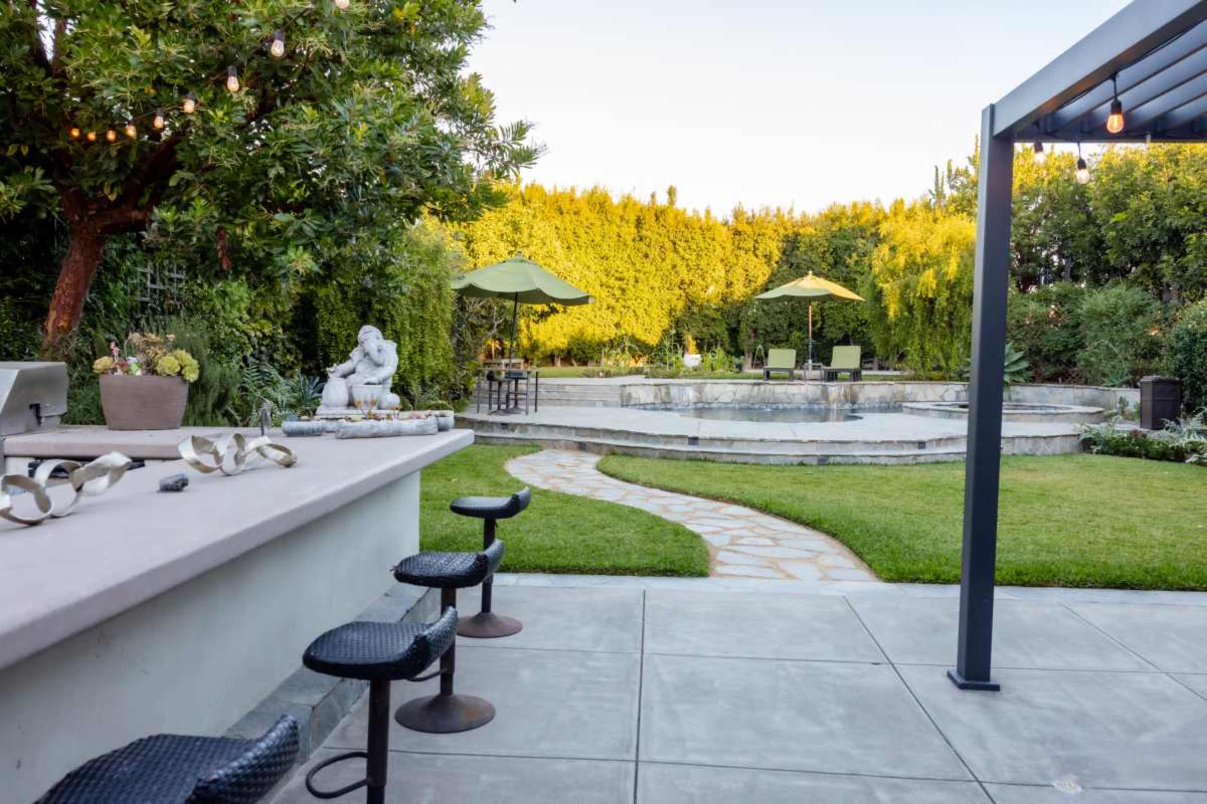 The image shows a landscaped backyard with a patio featuring bar stools, a stone pathway, and a seating area surrounded by greenery and umbrellas.