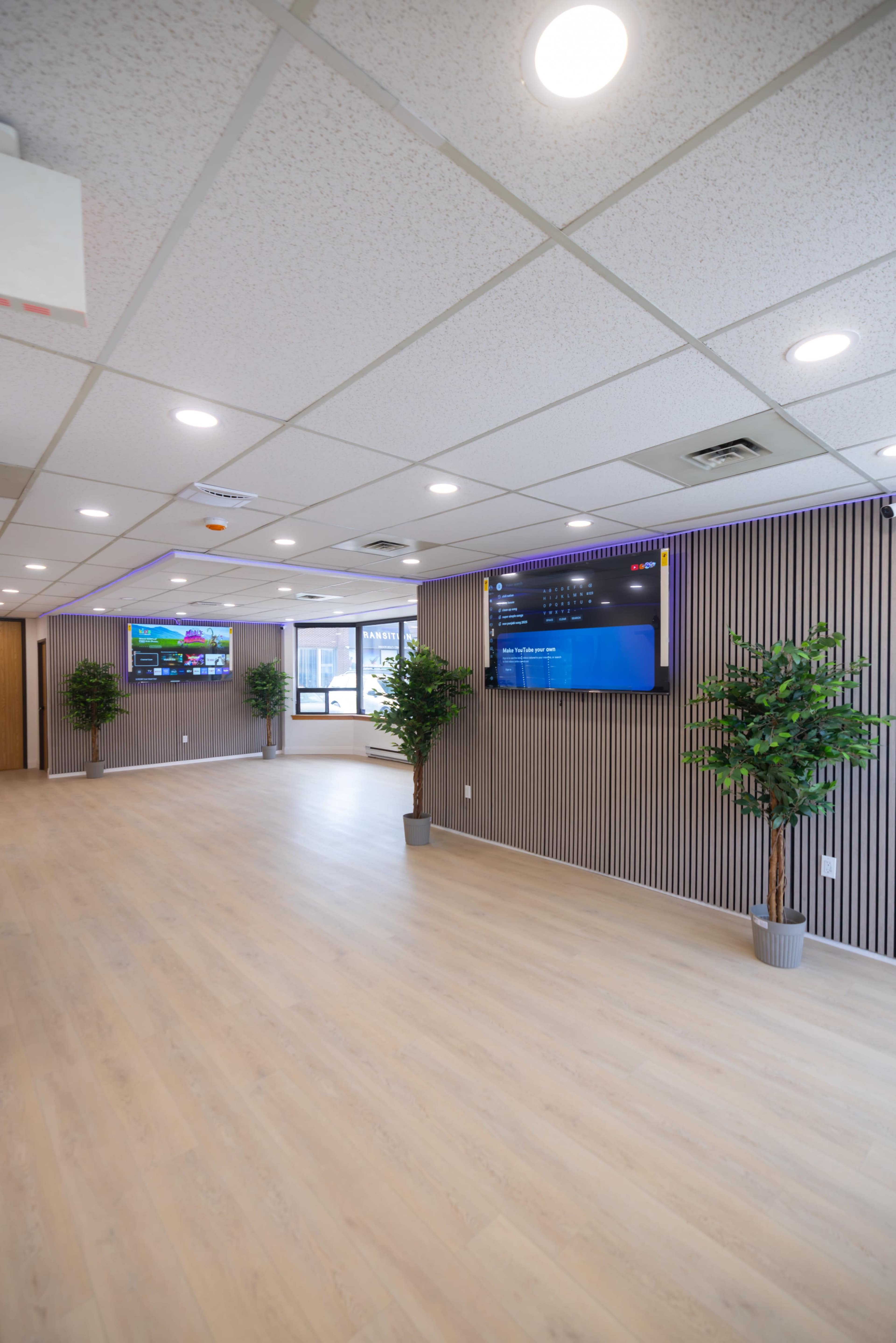 The image shows a spacious, well-lit indoor area featuring two wall-mounted screens and potted plants along the walls.