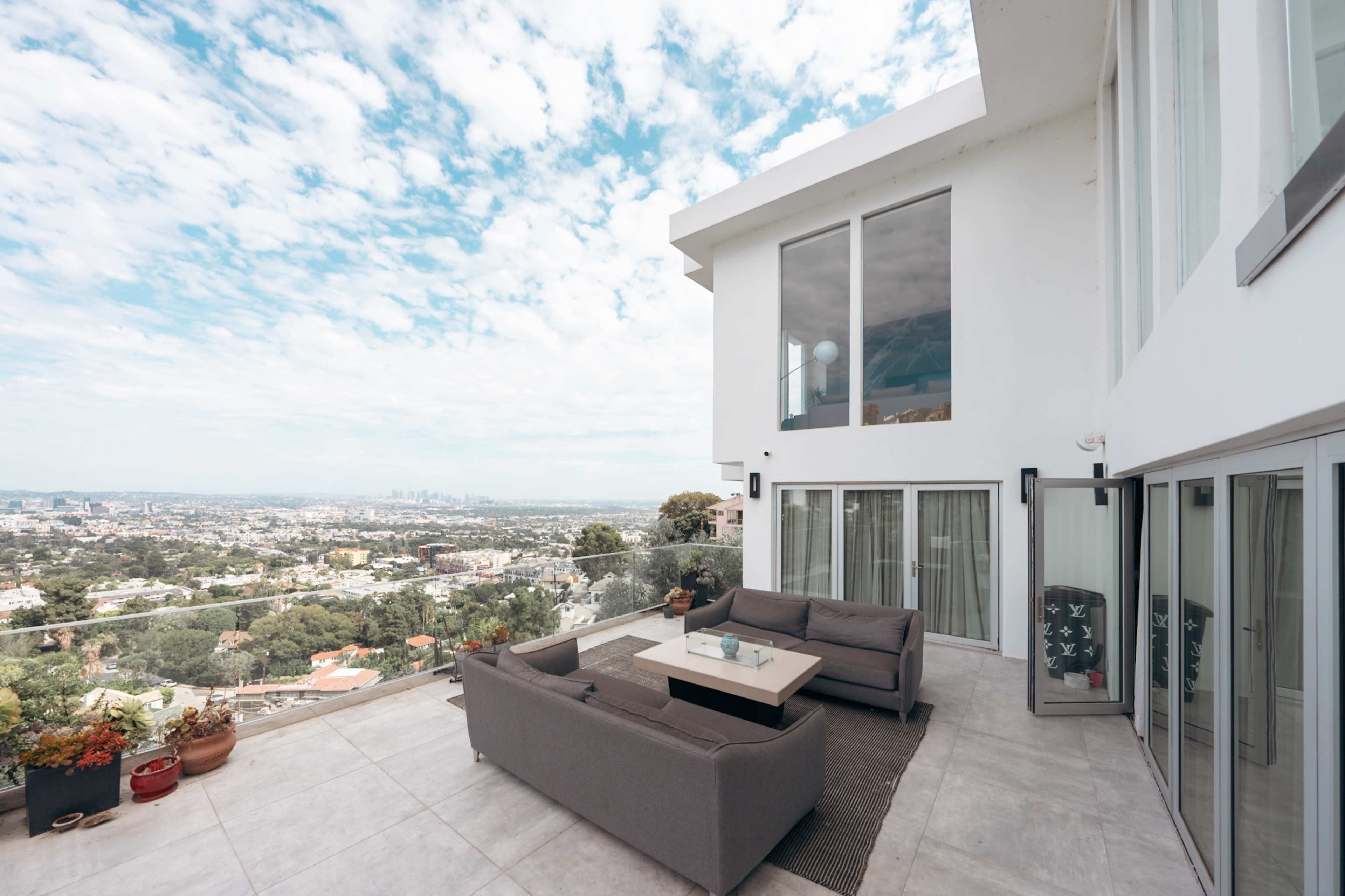 The image shows an outdoor patio area of a modern home, featuring a seating arrangement and a view of a cityscape under a partly cloudy sky.