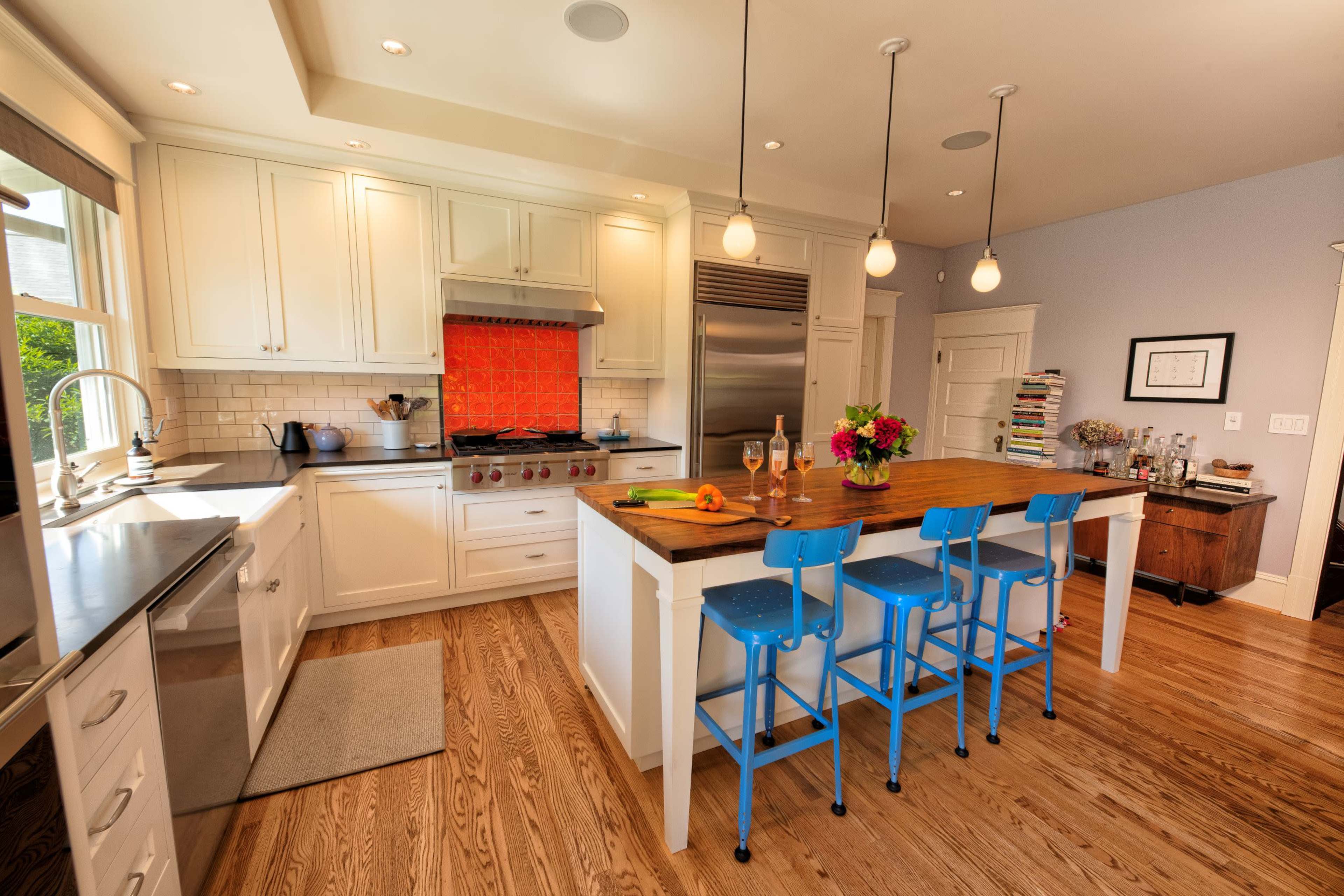 A modern kitchen features white cabinets, a large island with blue stools, and an orange tile backsplash behind the stove.