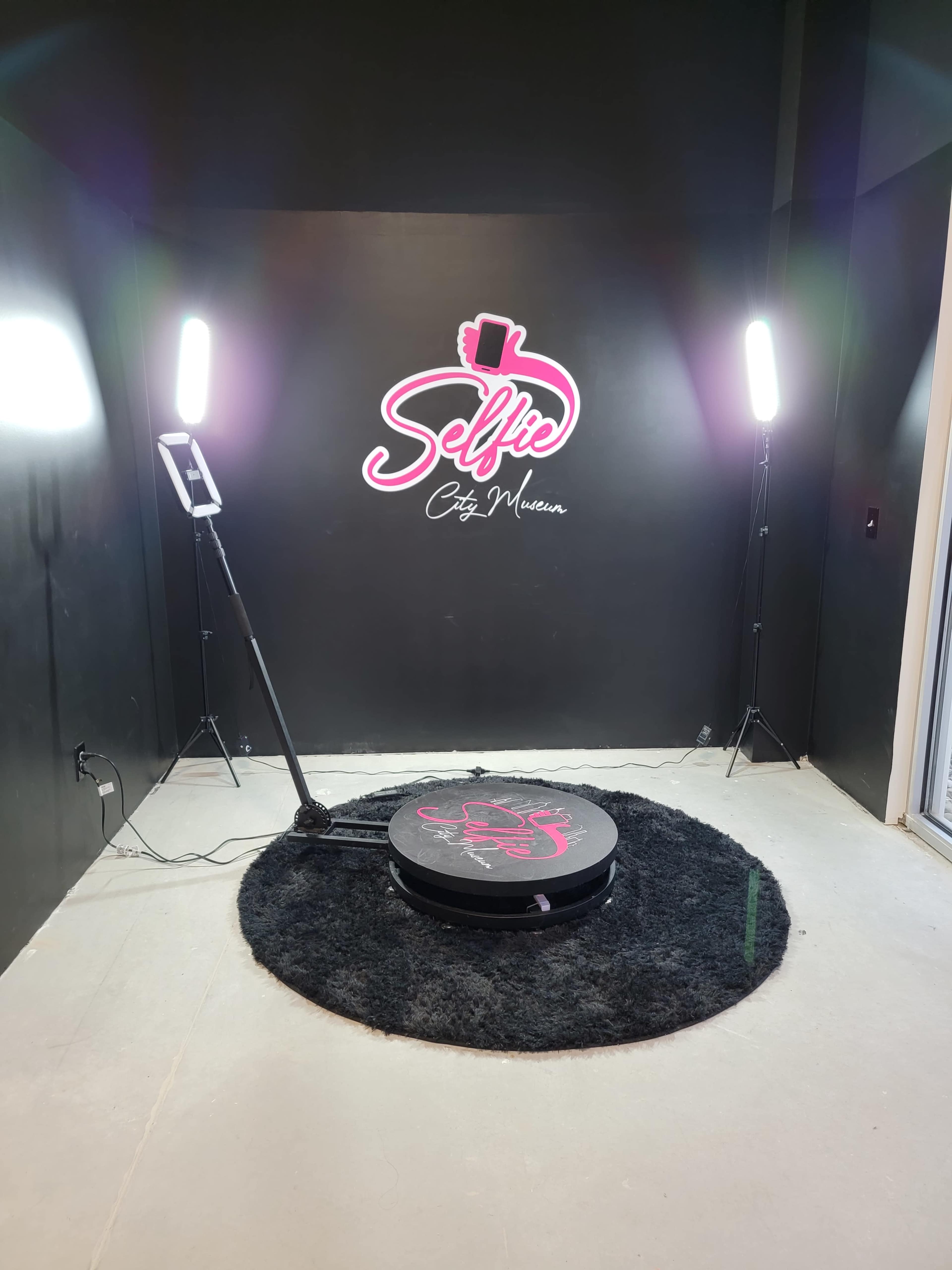 The image shows a photography setup for selfies, featuring a circular platform on a black rug, flanked by two bright lights, and a "Selfie" sign on a black wall.