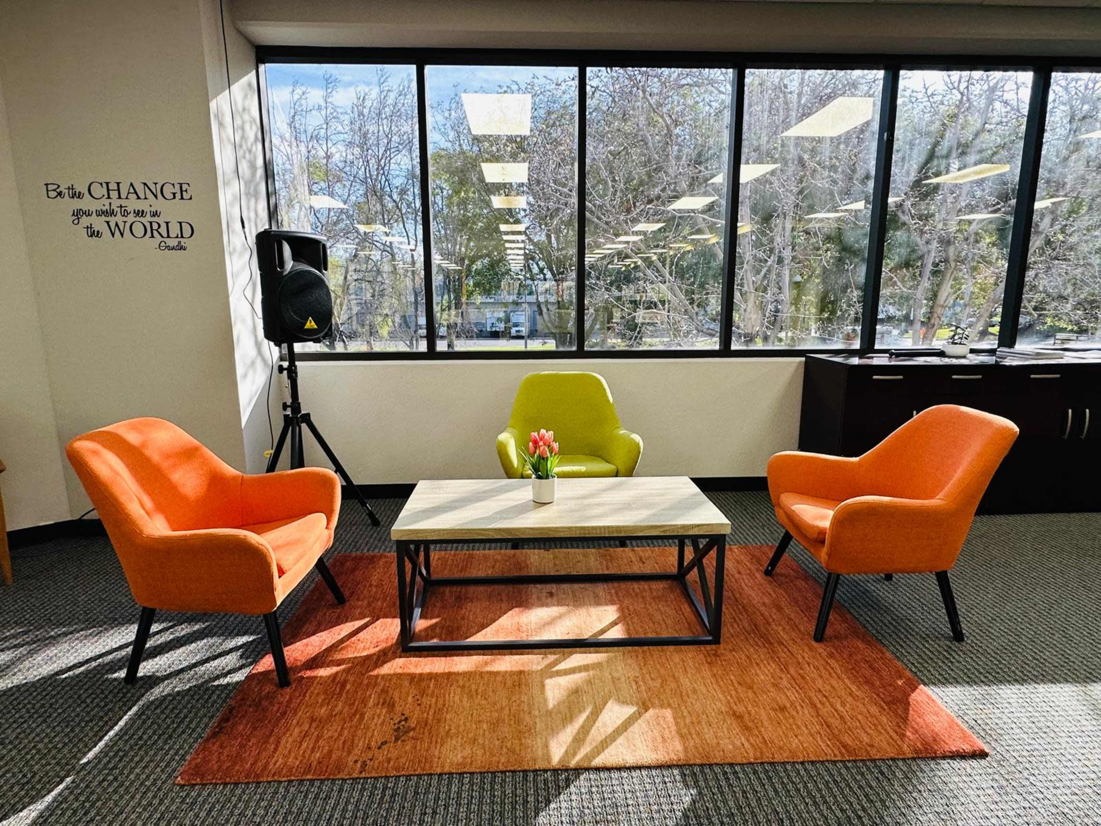 The image shows a modern office space with three colorful chairs surrounding a coffee table, placed on a textured rug near large windows.