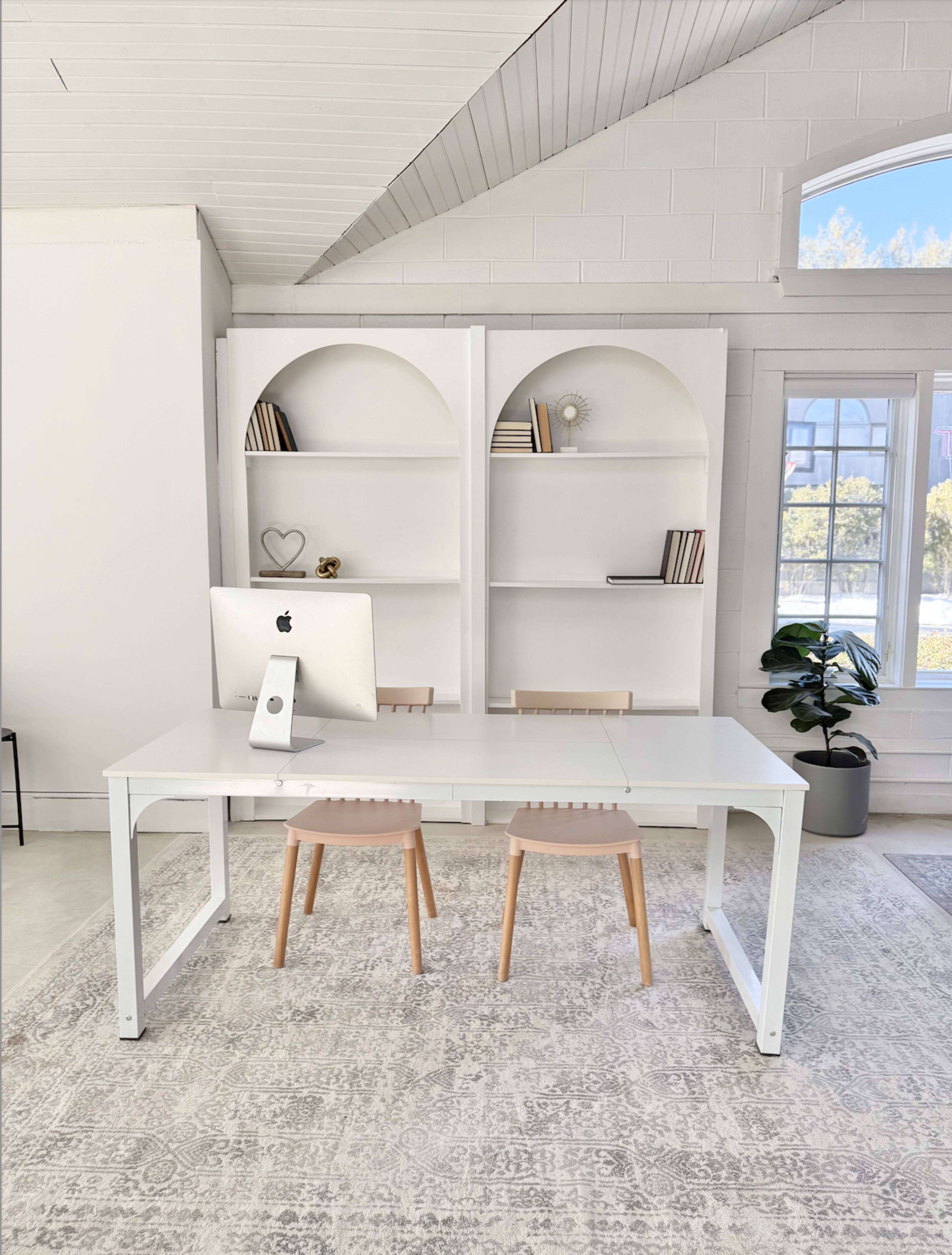 The image shows a minimalist workspace with a white desk, two wooden chairs, an iMac, and a bookshelf decorated with books and a small decorative item.