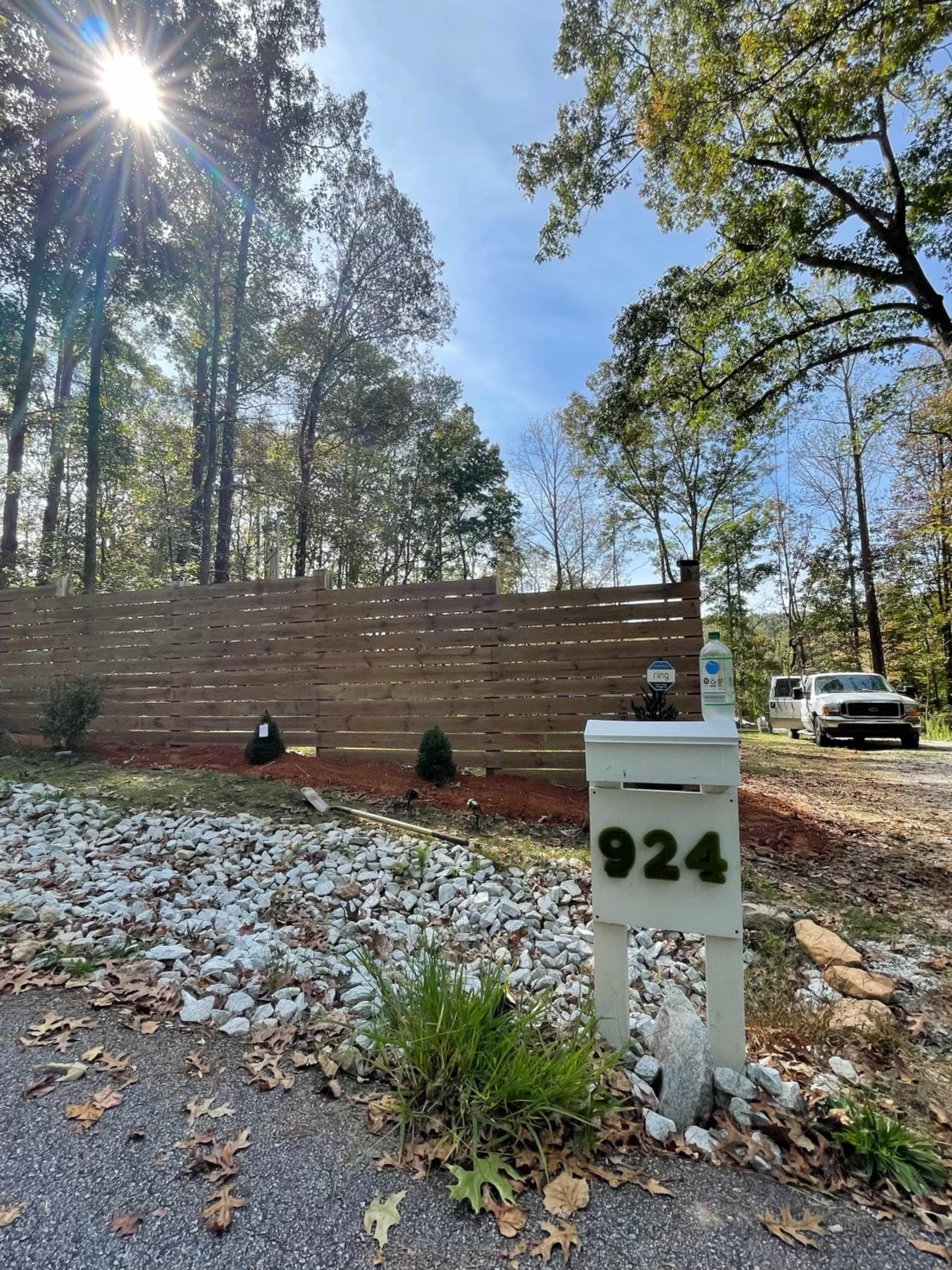 A white mailbox marked "924" stands near a gravel driveway, with a wooden fence and trees in the background under a clear blue sky.