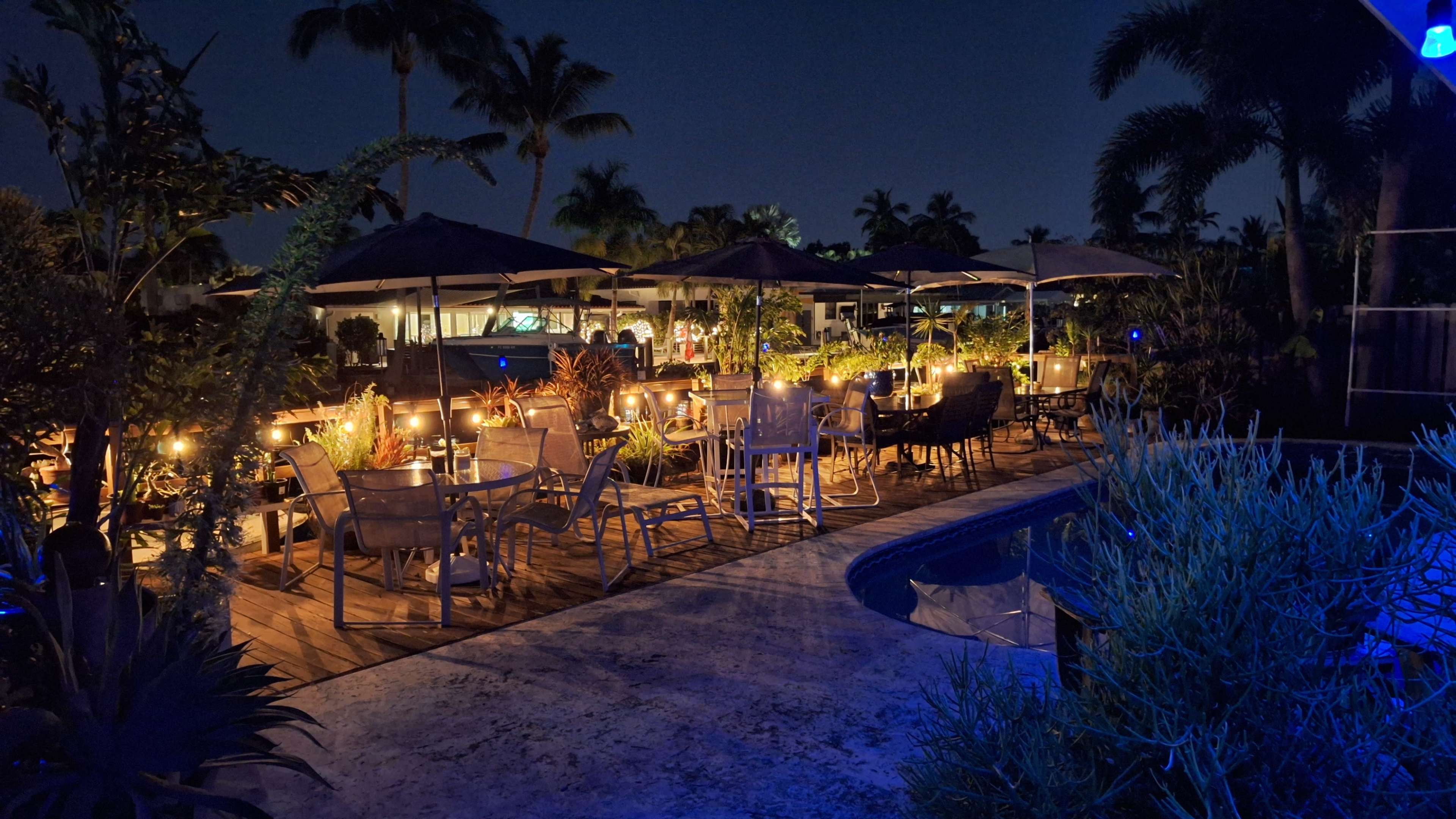The scene shows a tropical outdoor dining area at night, featuring illuminated tables and umbrellas surrounded by lush greenery and a swimming pool.