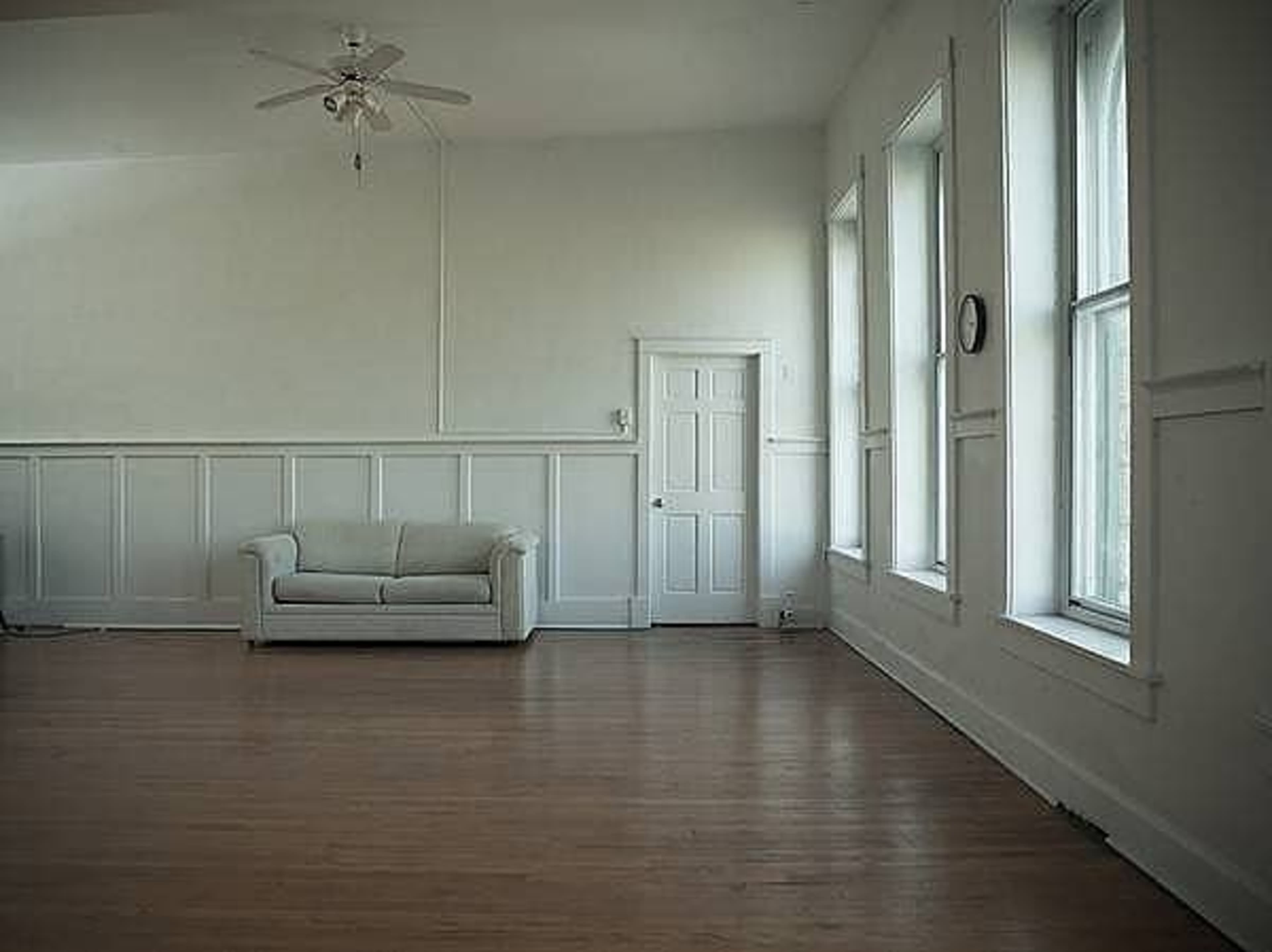 A spacious room with wooden flooring, white walls, a light-colored couch, and large windows.