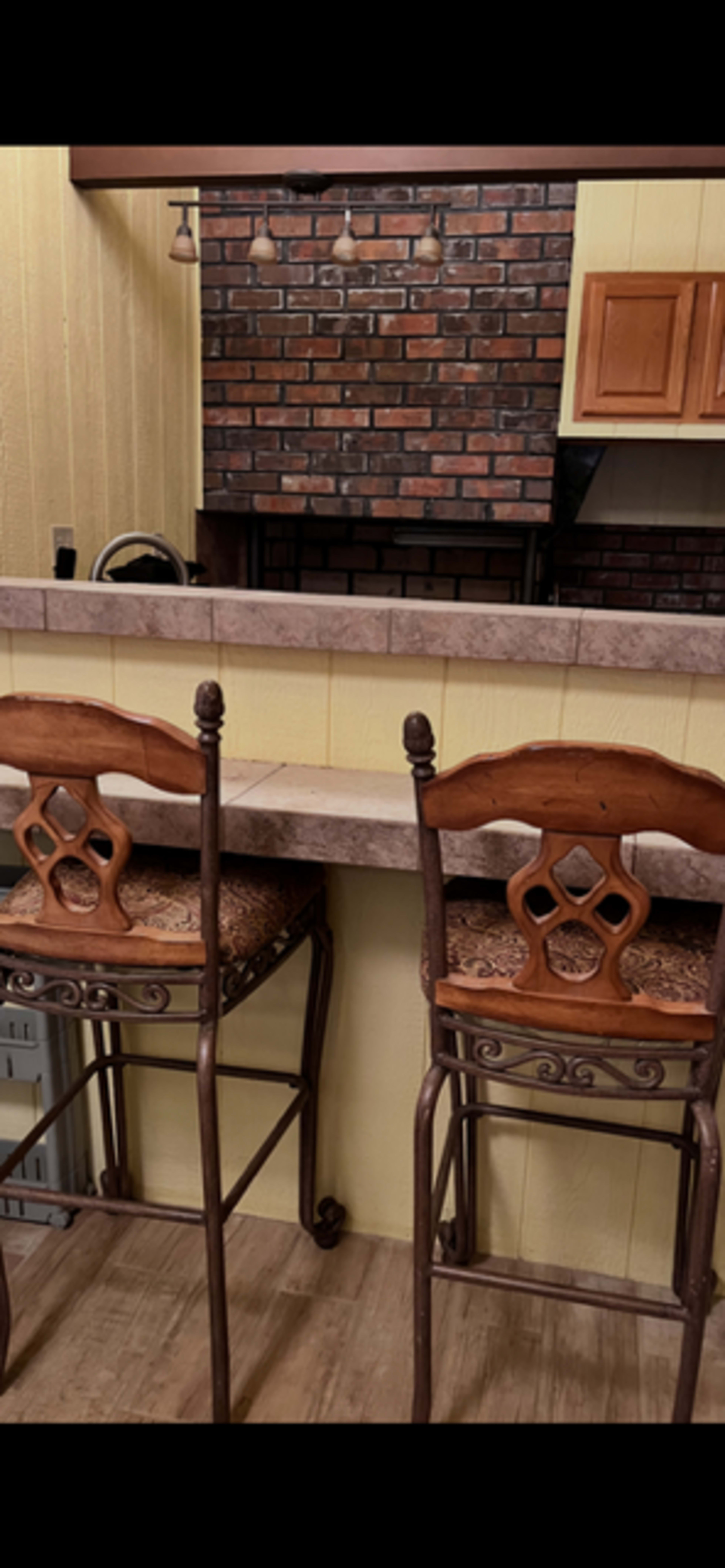 The image shows a bar area with two high wooden stools positioned in front of a countertop, set against a brick wall and a kitchen area in the background.
