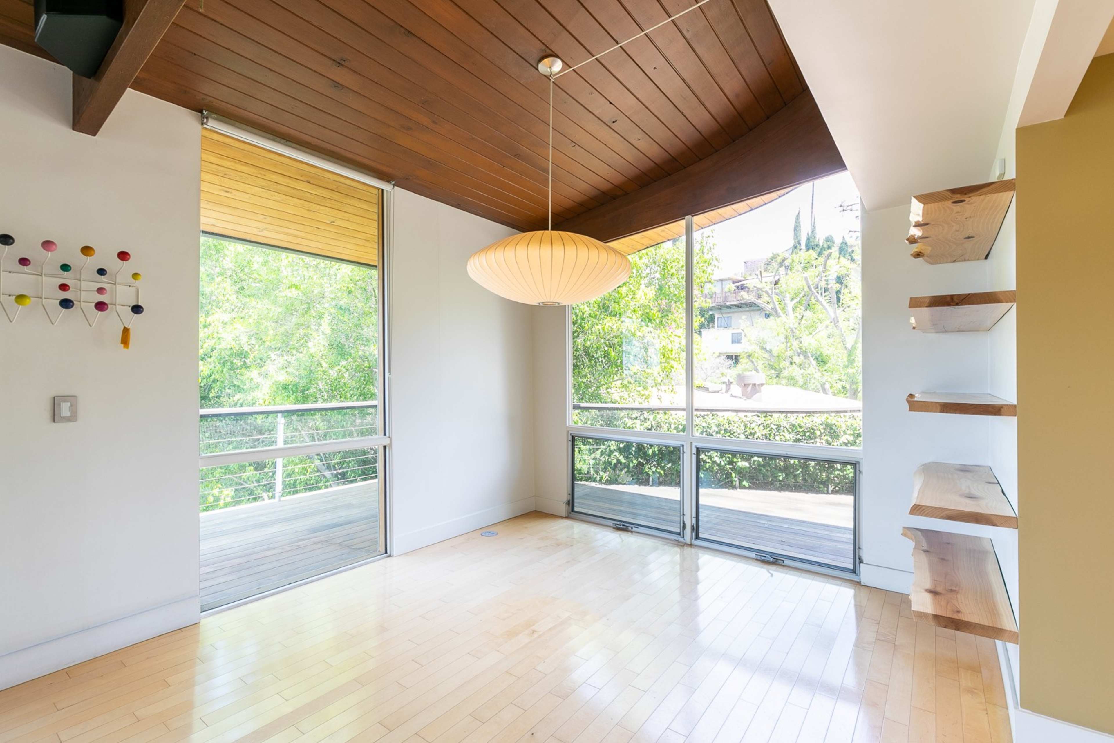 A spacious room features large windows overlooking greenery, a wooden ceiling, and minimalist shelving.