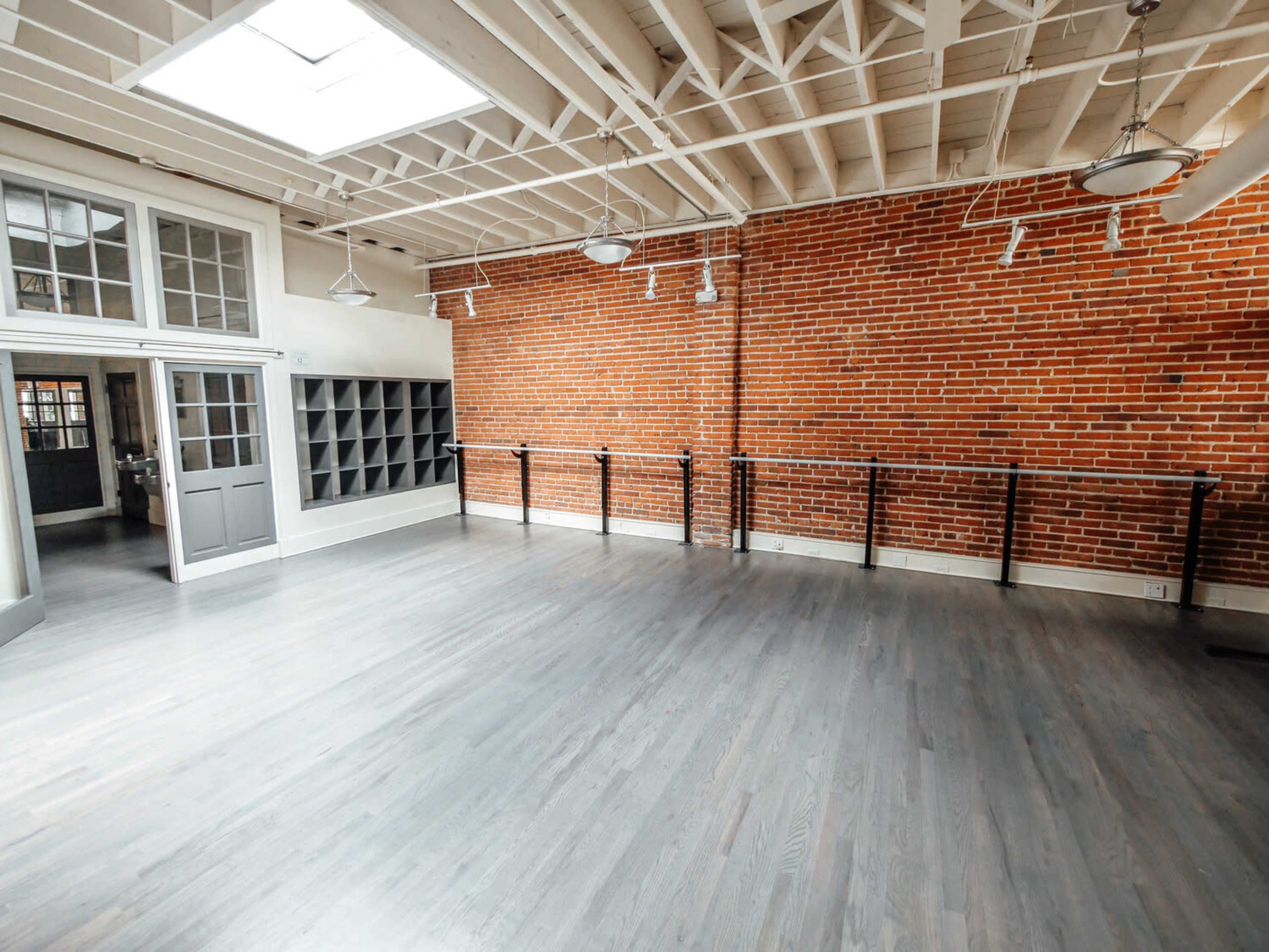 The image shows a spacious, well-lit dance studio with exposed brick walls, hardwood flooring, and ballet bars along one side.