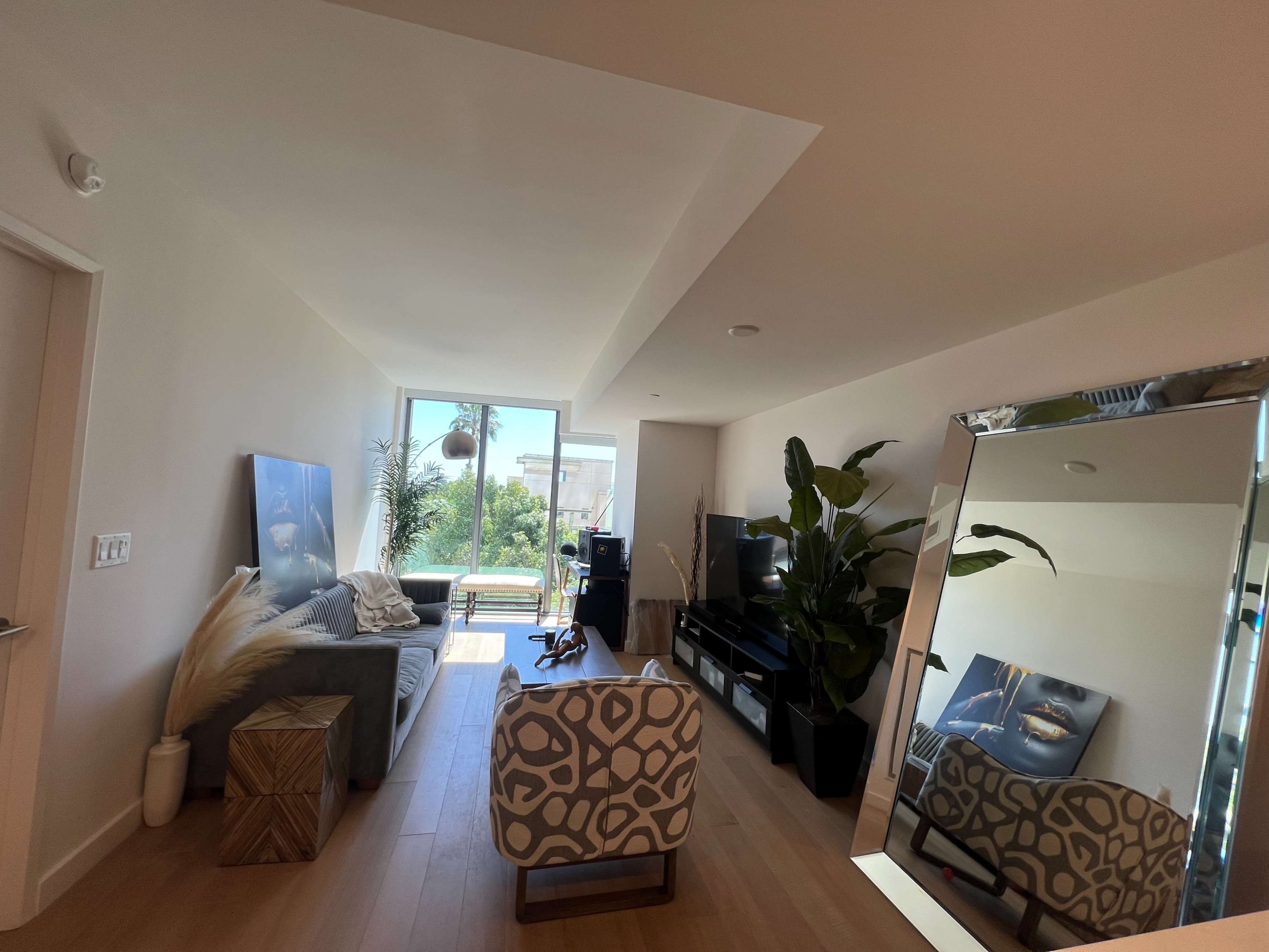 The image shows a modern living room featuring a sofa, a large mirror, a television, and a view of greenery through a window.