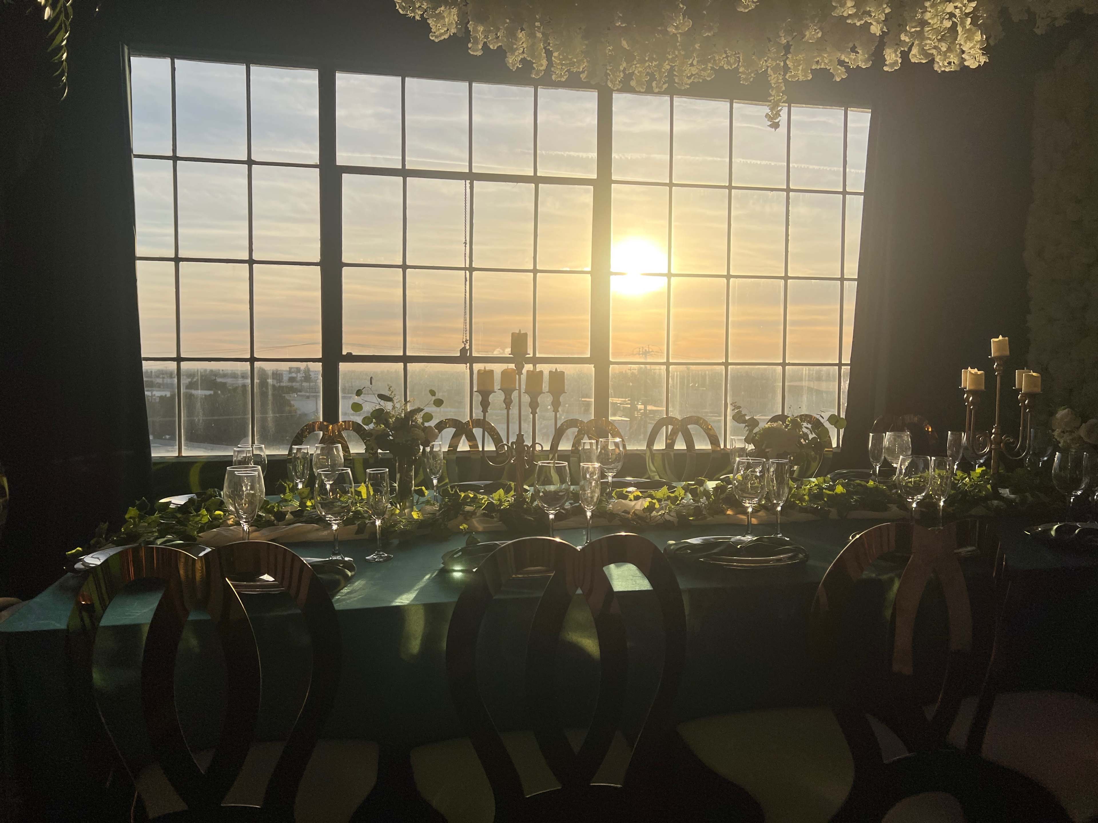 A beautifully arranged dining table featuring elegant table settings, with a backdrop of large windows displaying a sunset over a scenic landscape.