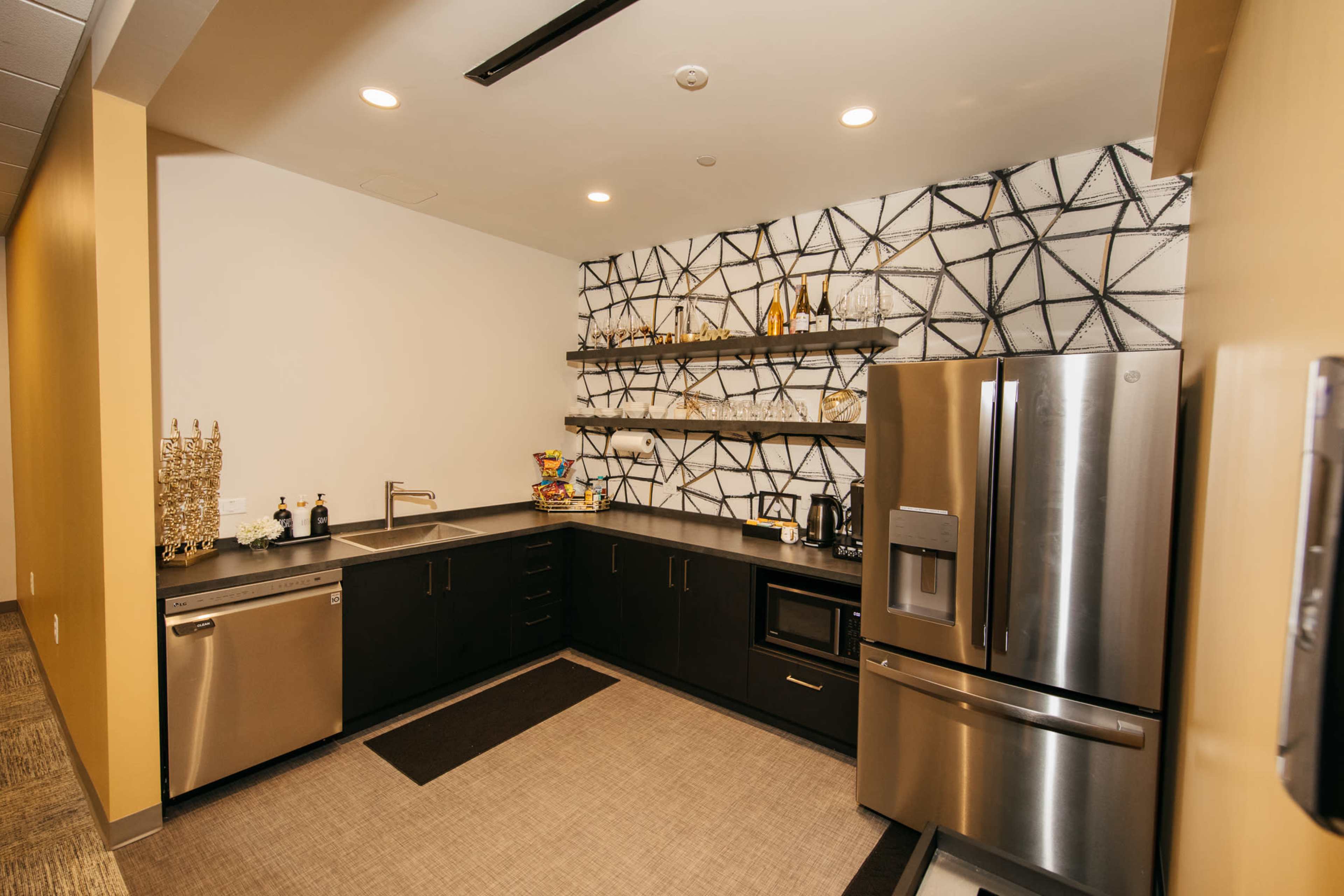 The image shows a modern kitchenette featuring dark cabinetry, a stainless steel refrigerator, and geometric-patterned wall shelves with various kitchen items.