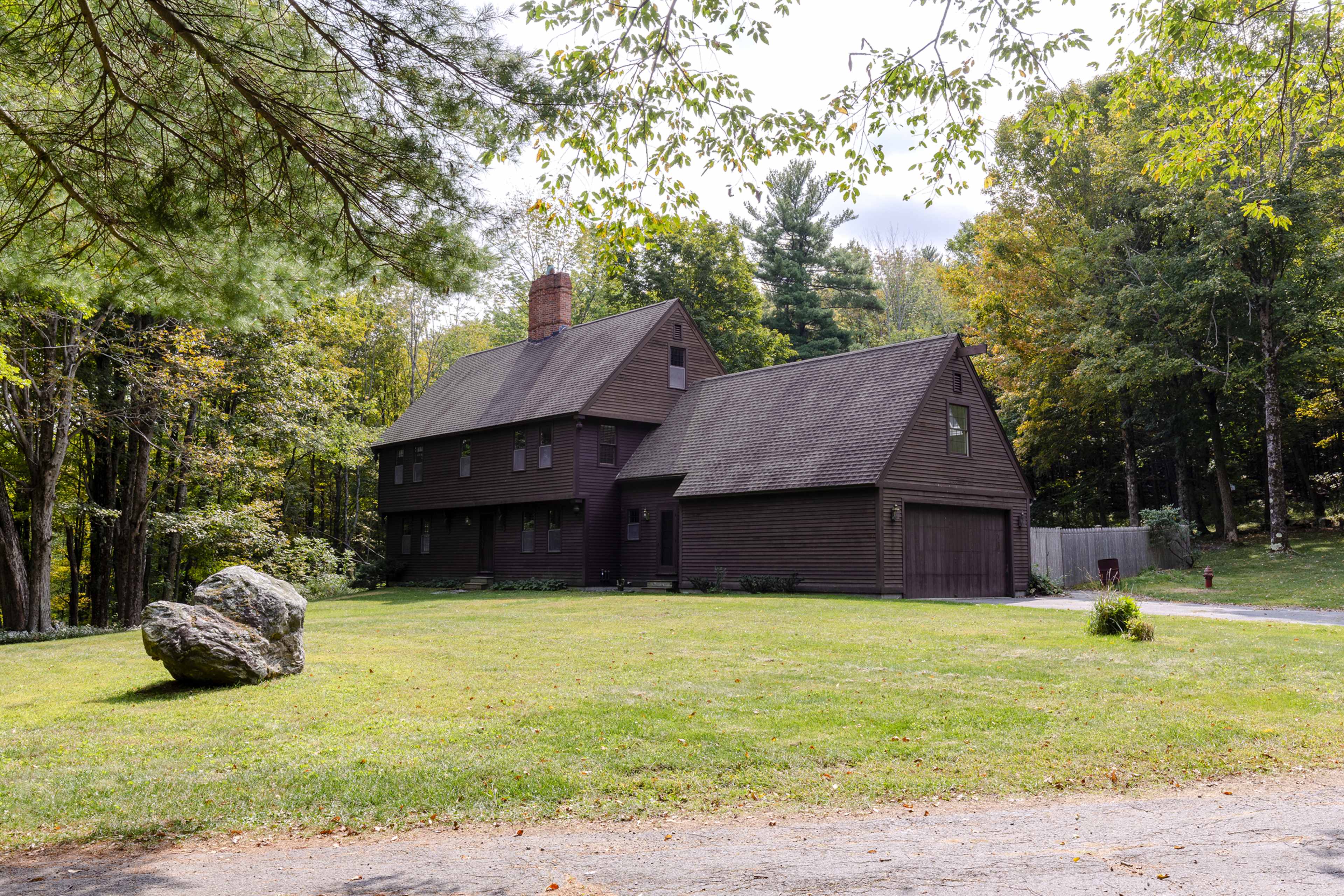 A large brown wooden house stands in a grassy area surrounded by trees, with a gravel driveway leading up to it.