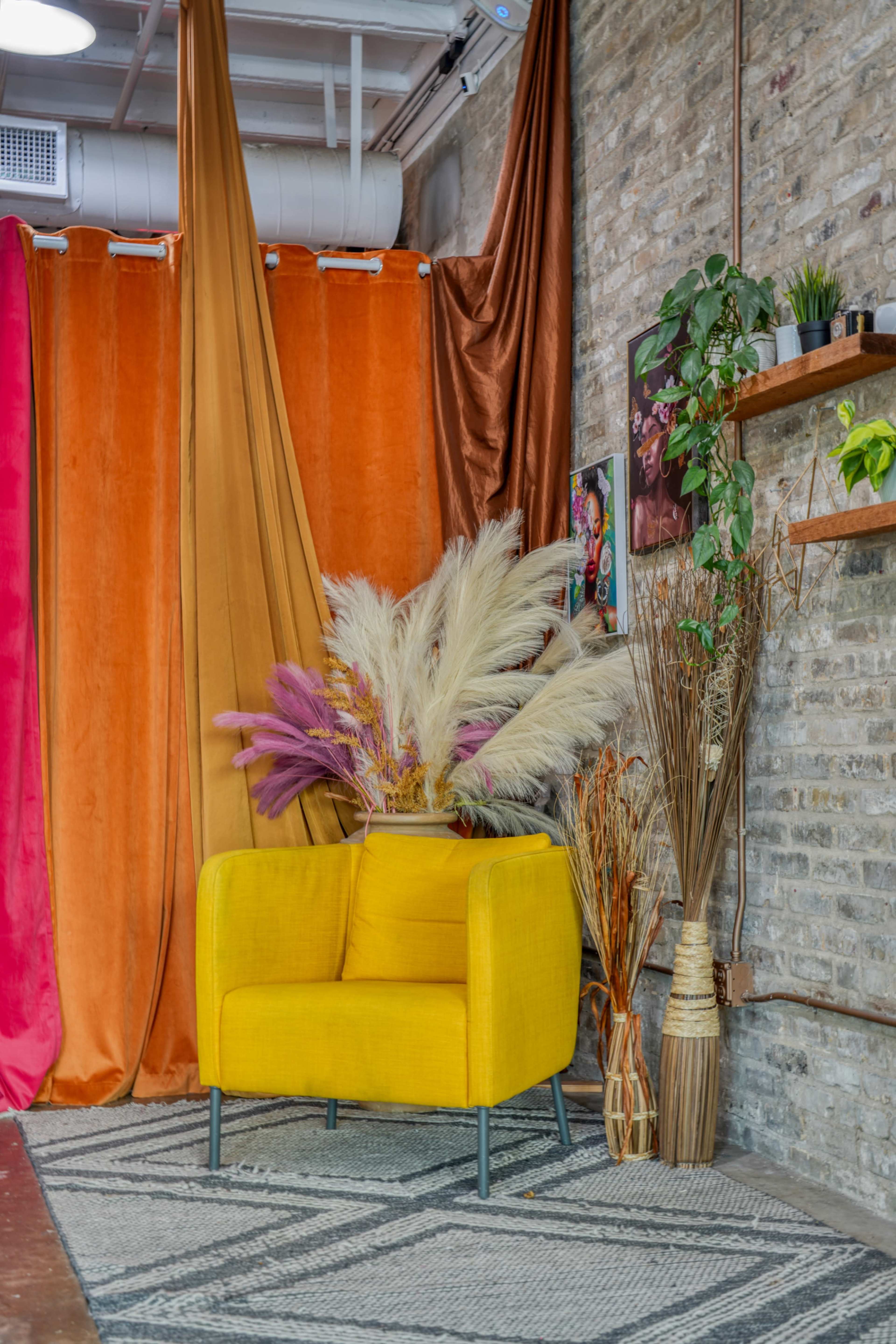 A yellow chair is positioned in front of colorful curtains and decorative plants against a brick wall.