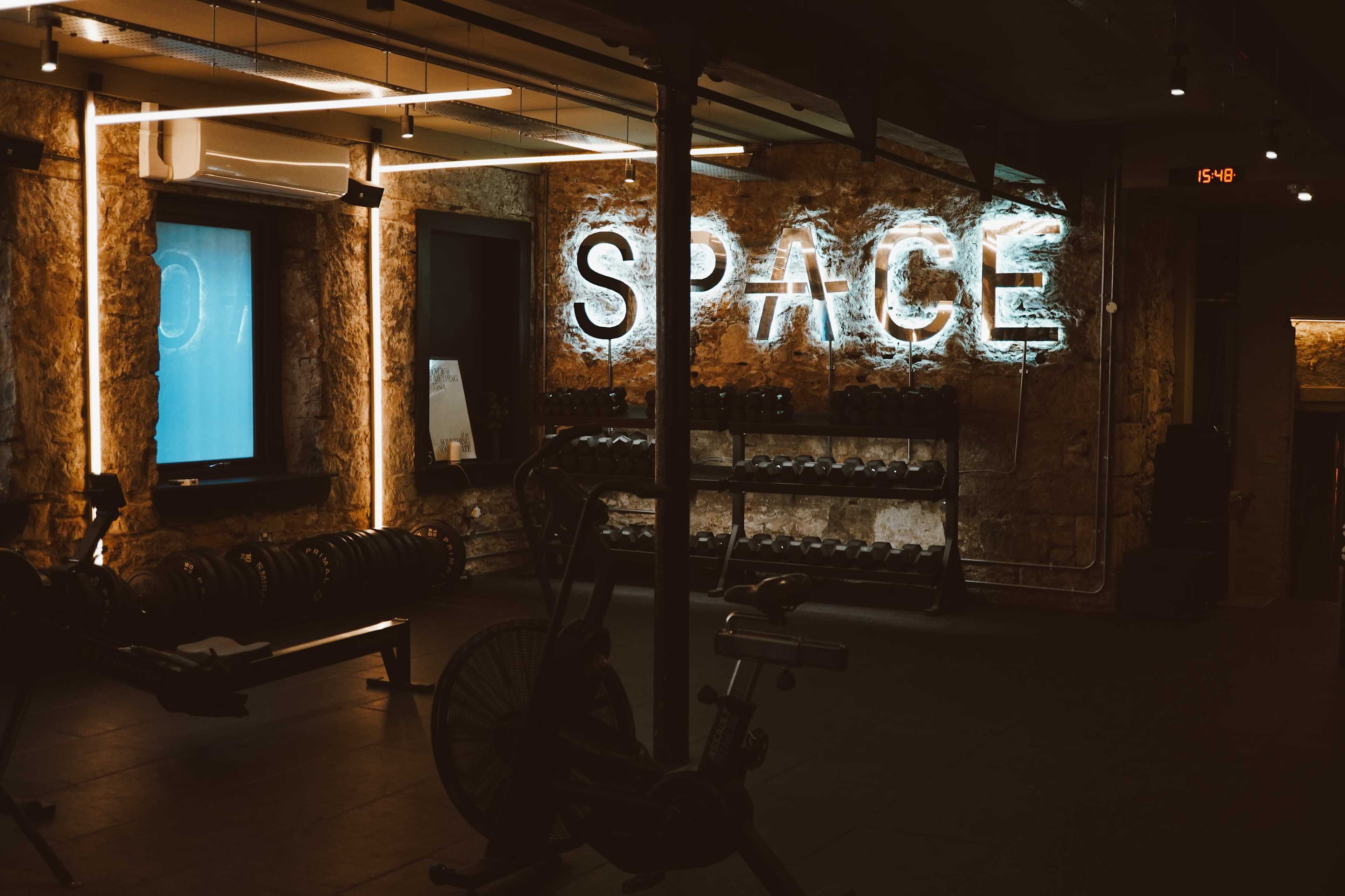 The image shows a gym interior featuring a wall with the word "SPACE" illuminated by neon lights, along with various gym equipment and free weights.