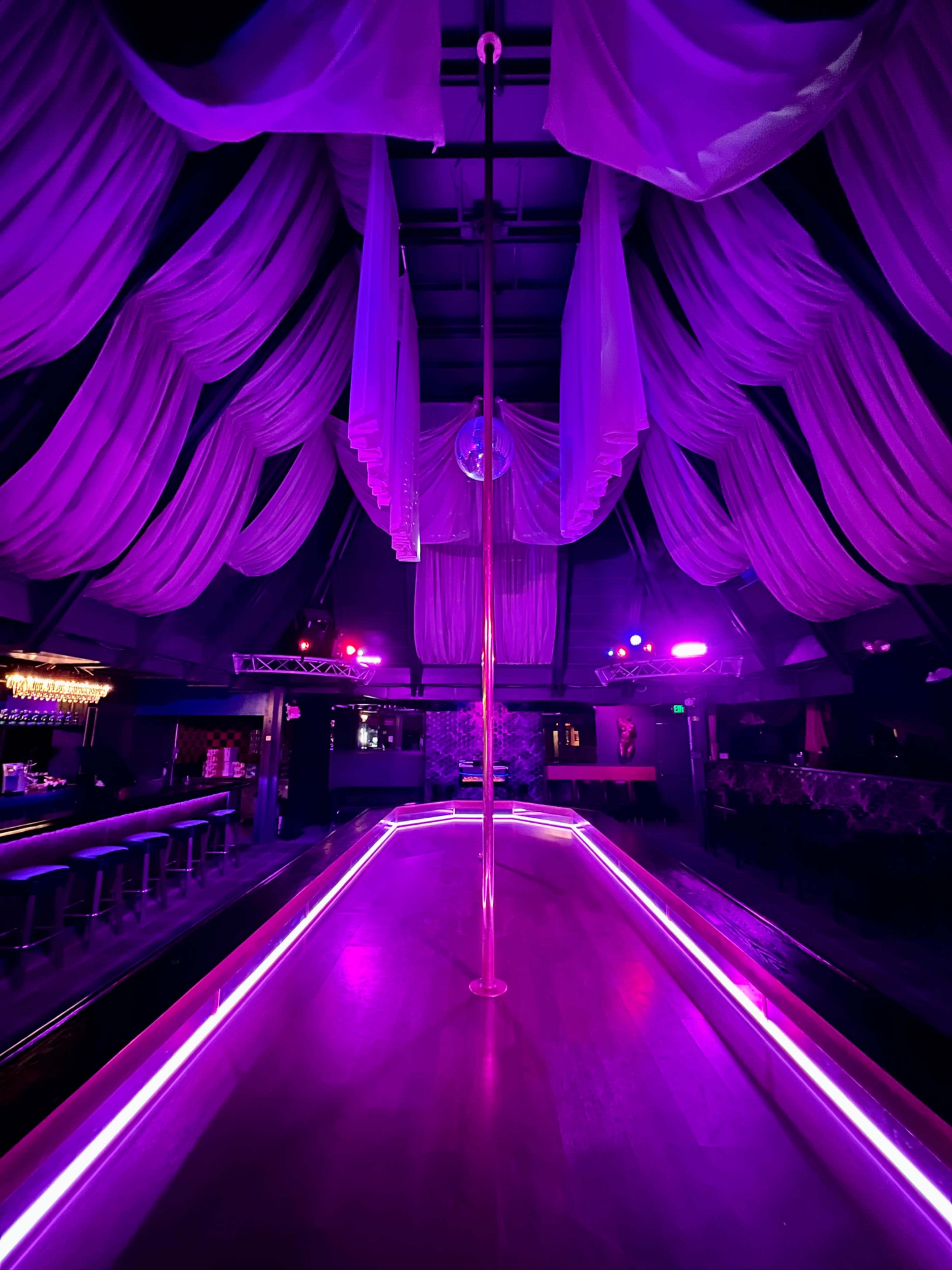 The interior of a dimly lit nightclub features a central pole surrounded by purple lighting and draped fabric overhead.