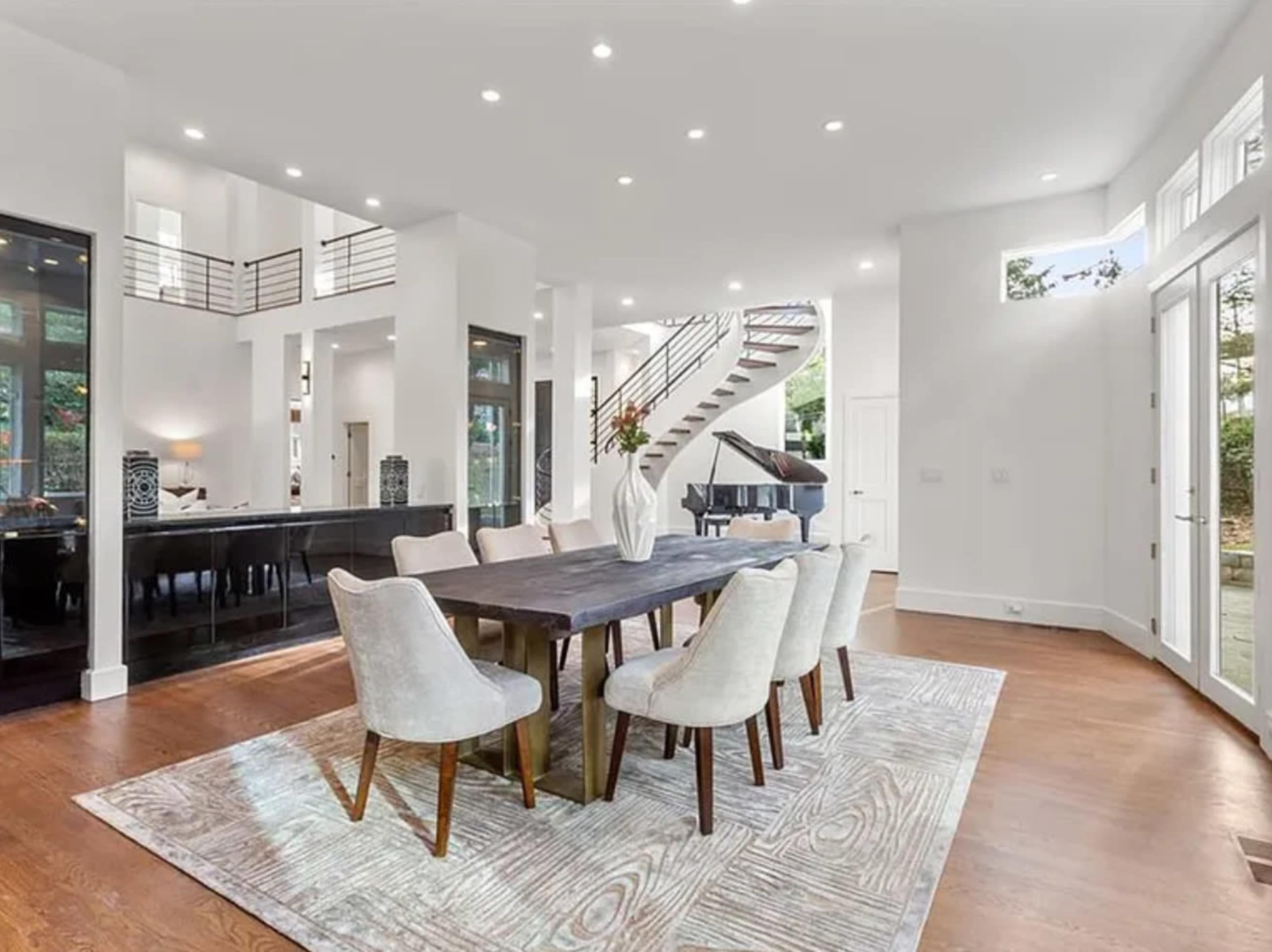 A modern dining area features a large wooden table surrounded by six upholstered chairs, set against a backdrop of an open-plan space with large windows and a staircase.