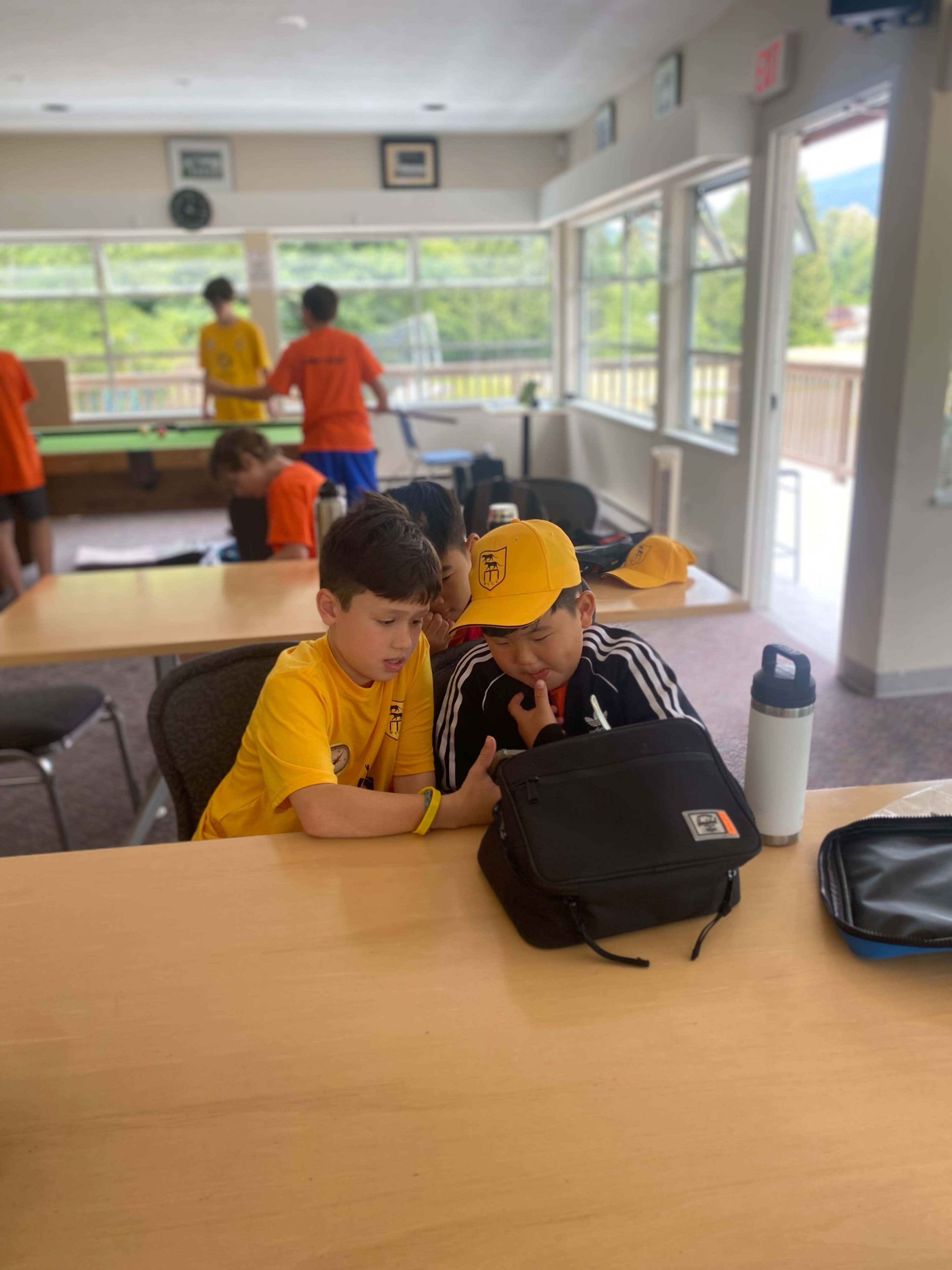 Two boys wearing yellow and black clothing sit closely together at a table, focusing on a mobile device while other children are visible in the background.
