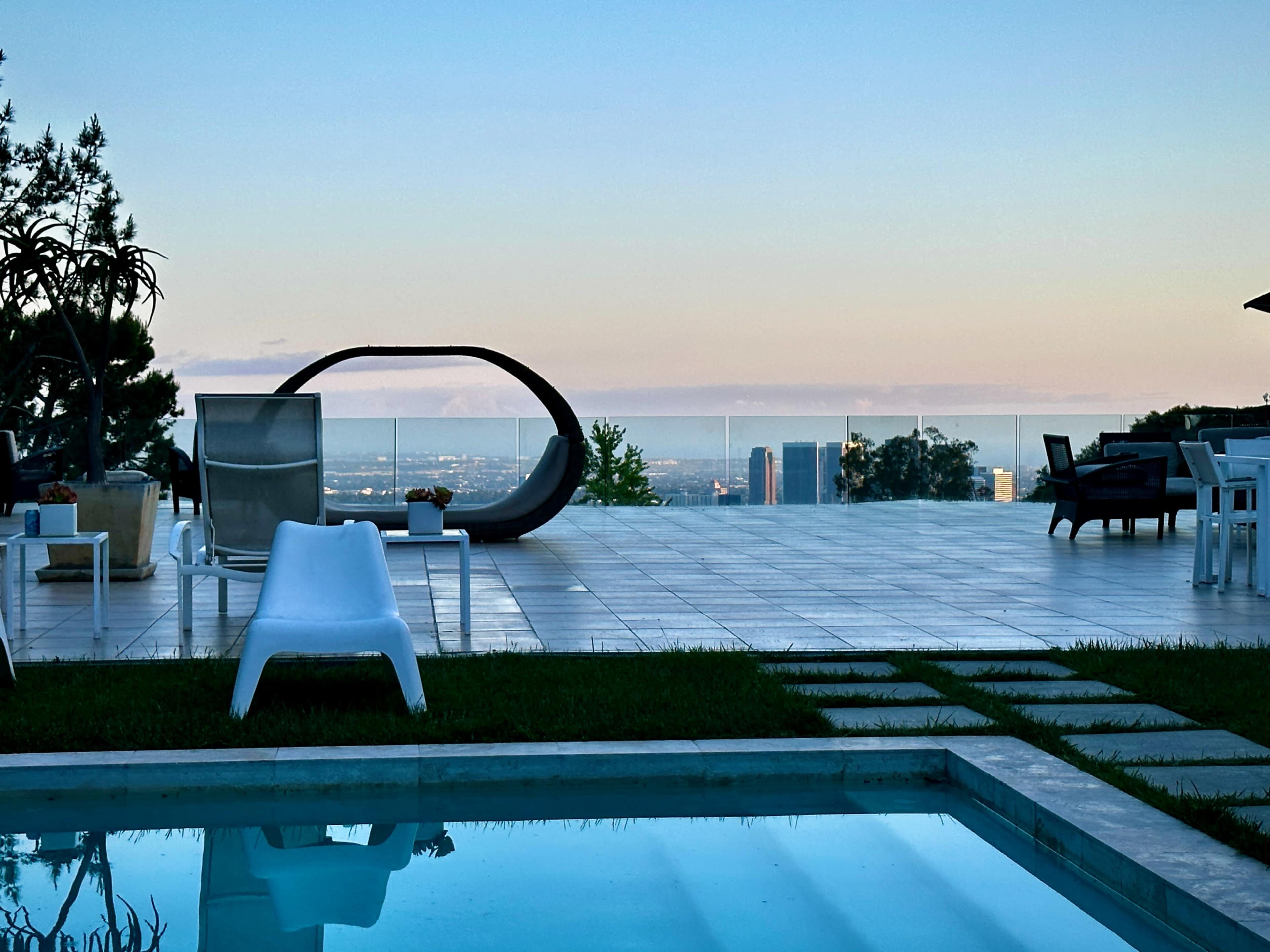 The image shows a modern outdoor area with a swimming pool, lounge chairs, and a scenic view of the city skyline at dusk.