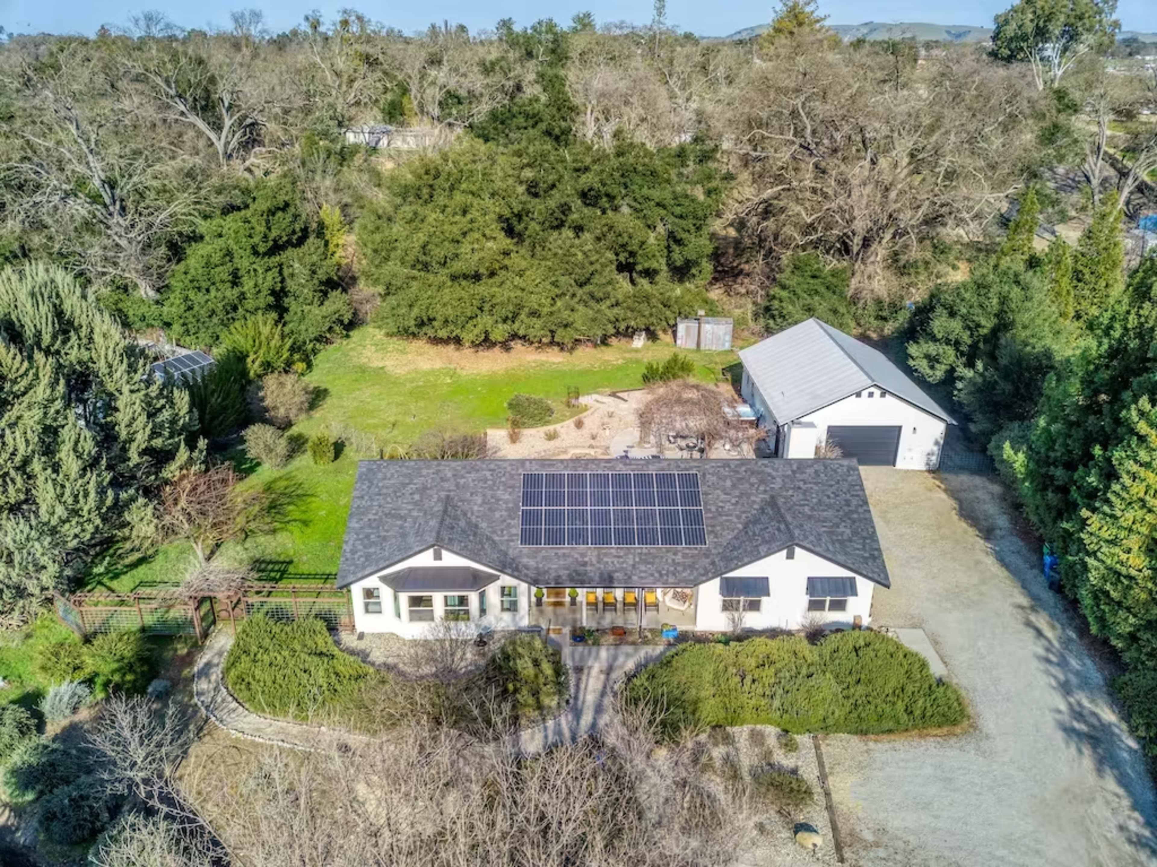 A single-story house with a solar panel roof is situated on a spacious lot surrounded by trees and a detached garage.