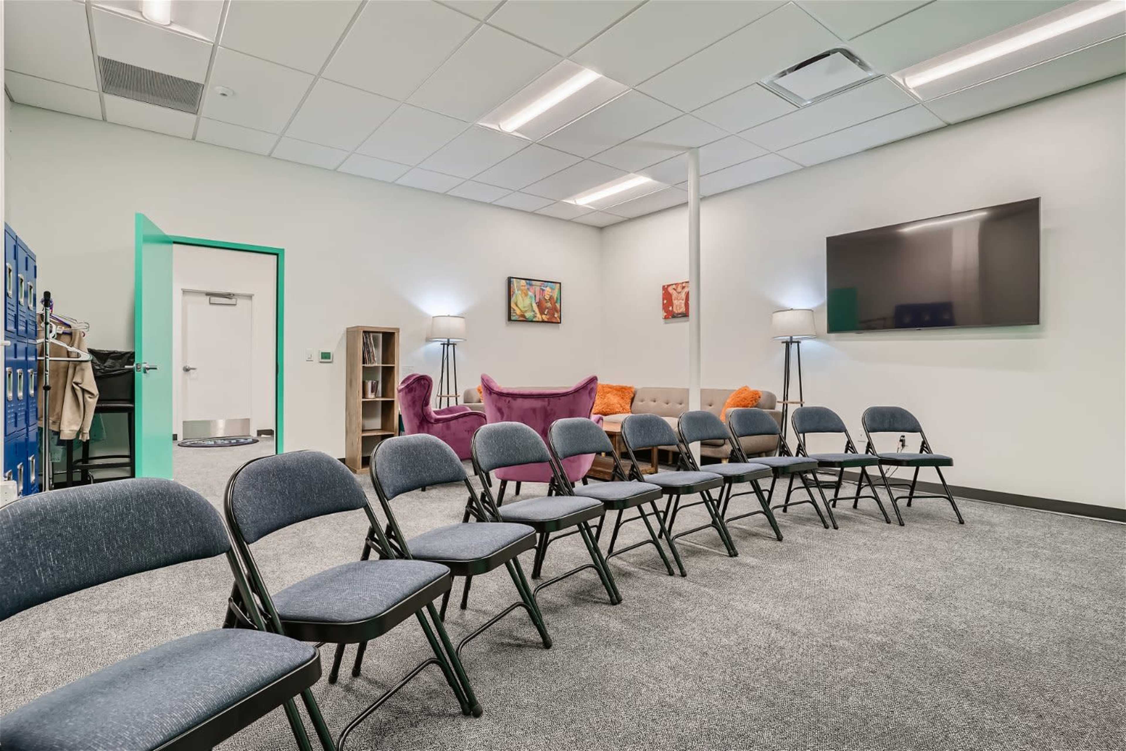 A room with a carpeted floor, featuring rows of folding chairs facing a wall-mounted TV, along with a couch, armchairs, and lockers in the background.
