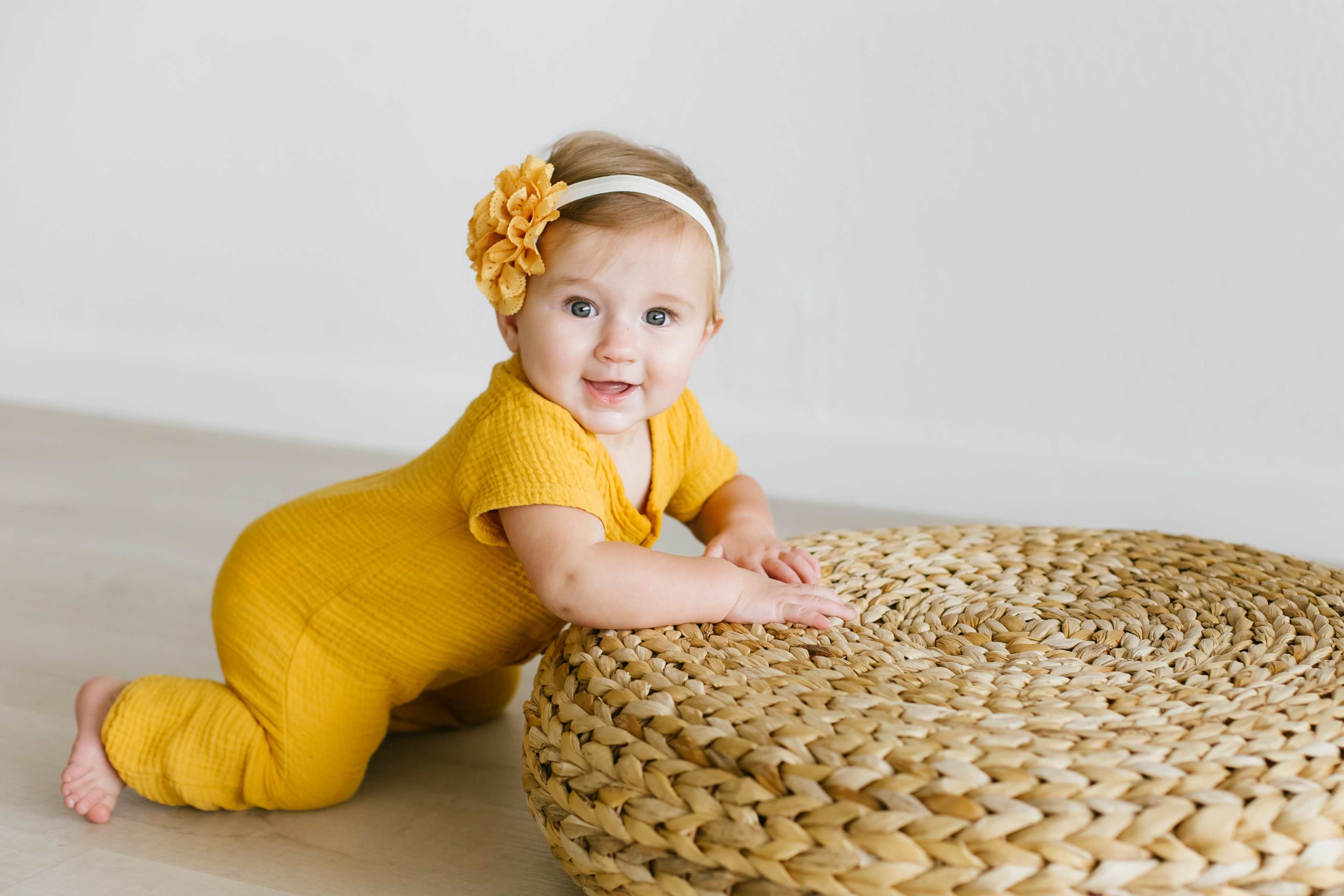 A baby in a yellow outfit is kneeling beside a large woven basket.