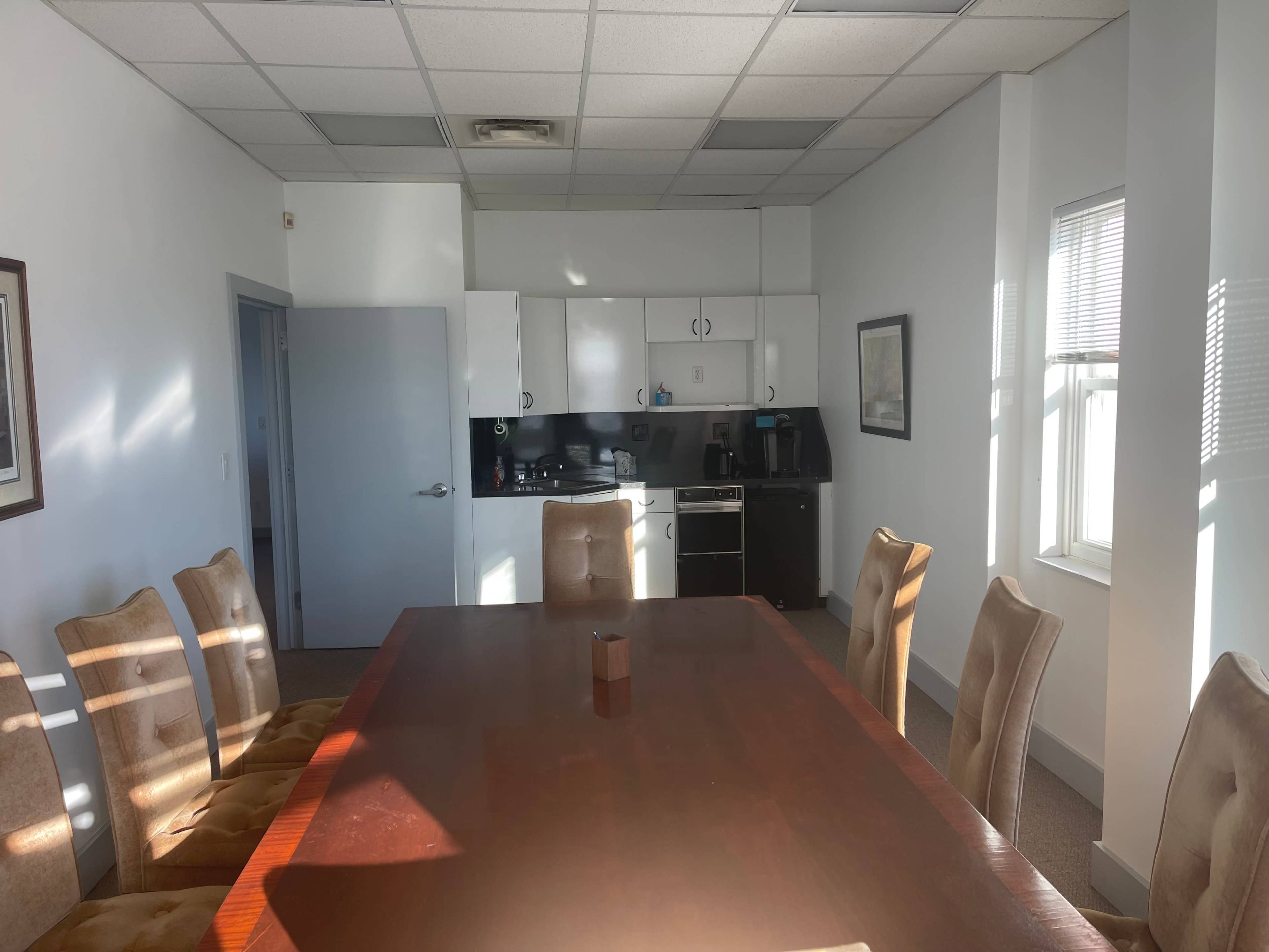 A conference room with a large wooden table surrounded by upholstered chairs, adjacent to a kitchenette with appliances and a window letting in natural light.