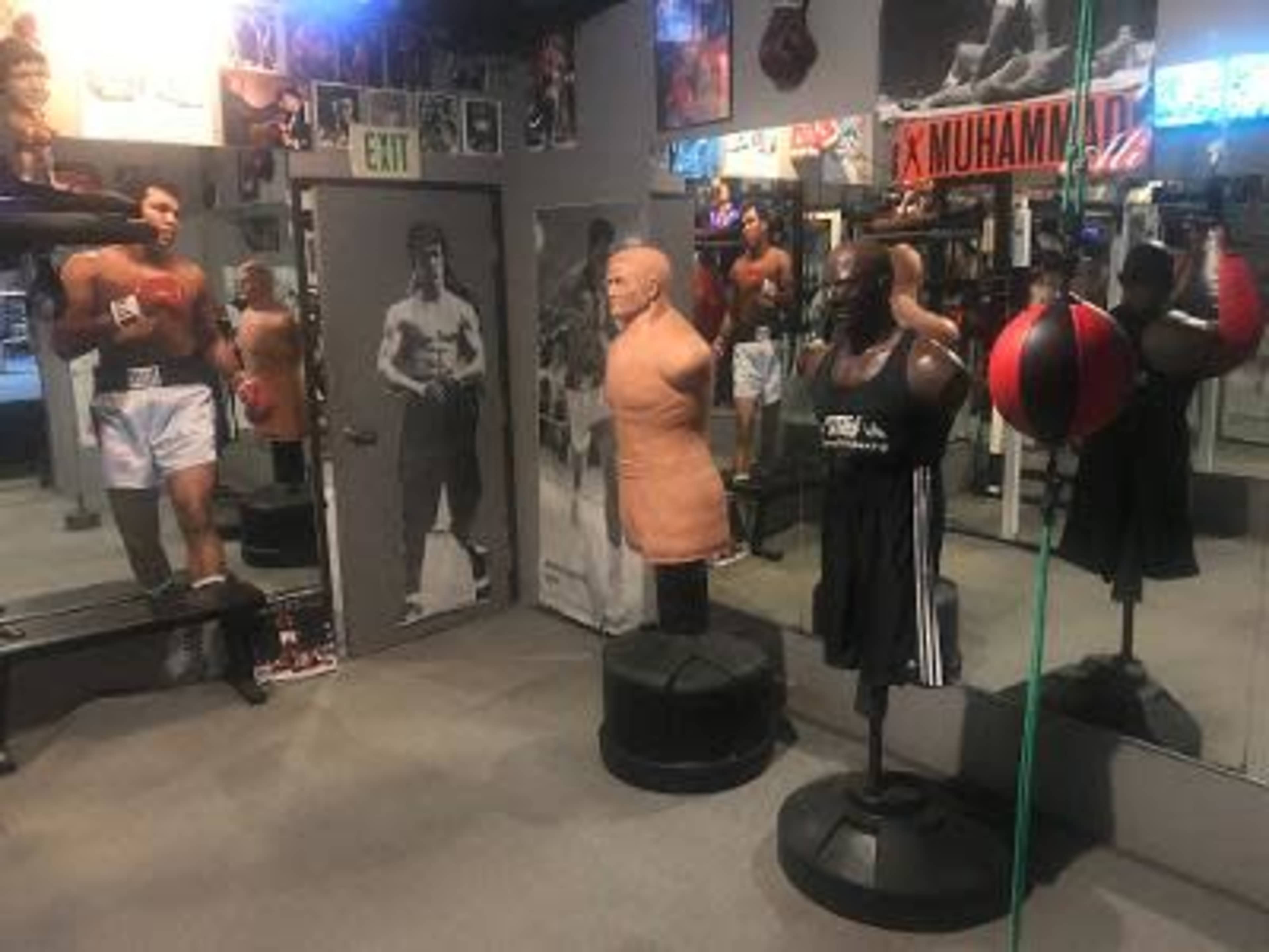 The image shows a boxing gym featuring a training area with mannequins, a heavy bag, and photos of renowned boxers on the walls.