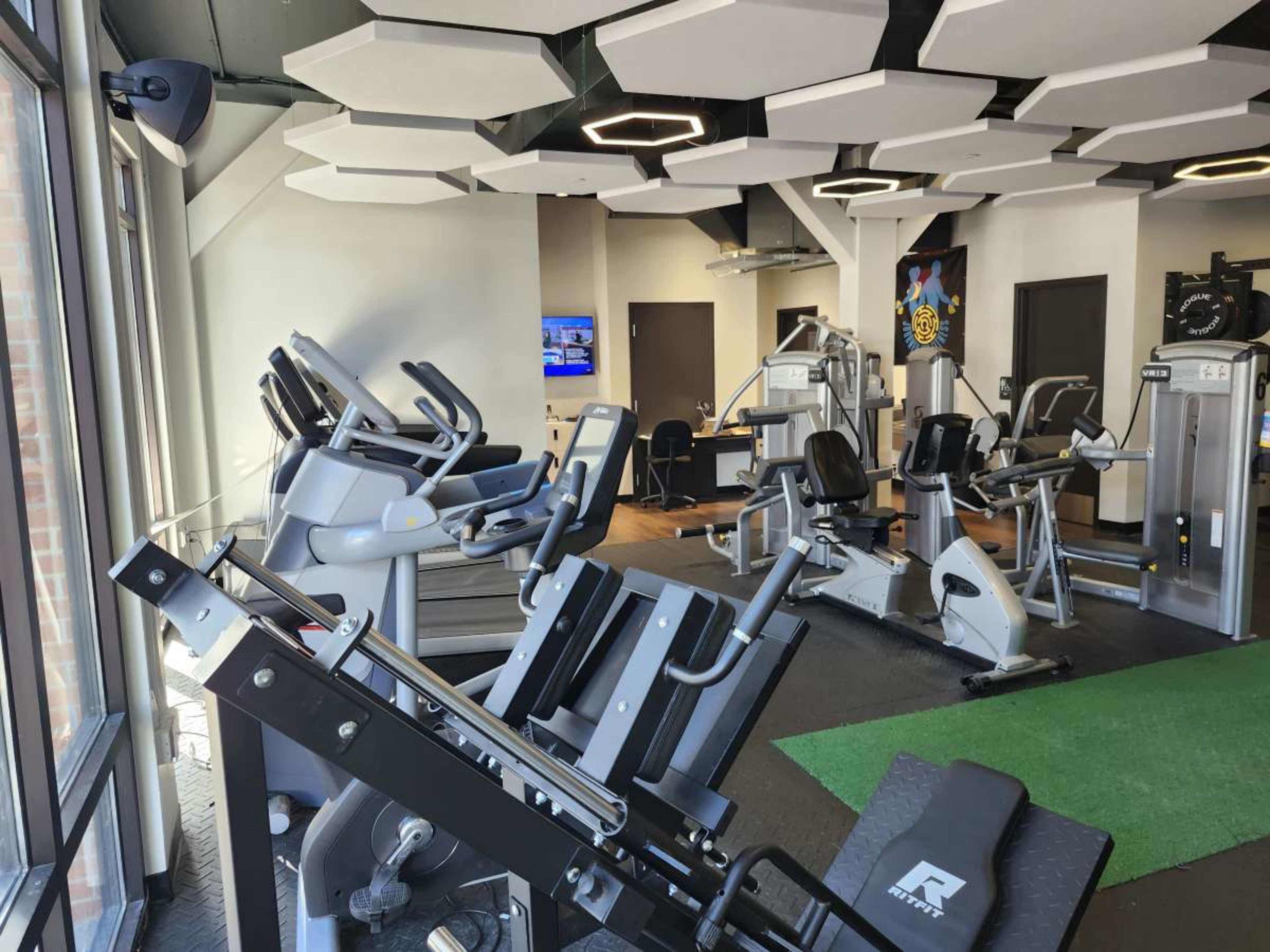 The image shows a modern gym interior featuring various exercise machines and equipment with large windows allowing natural light.