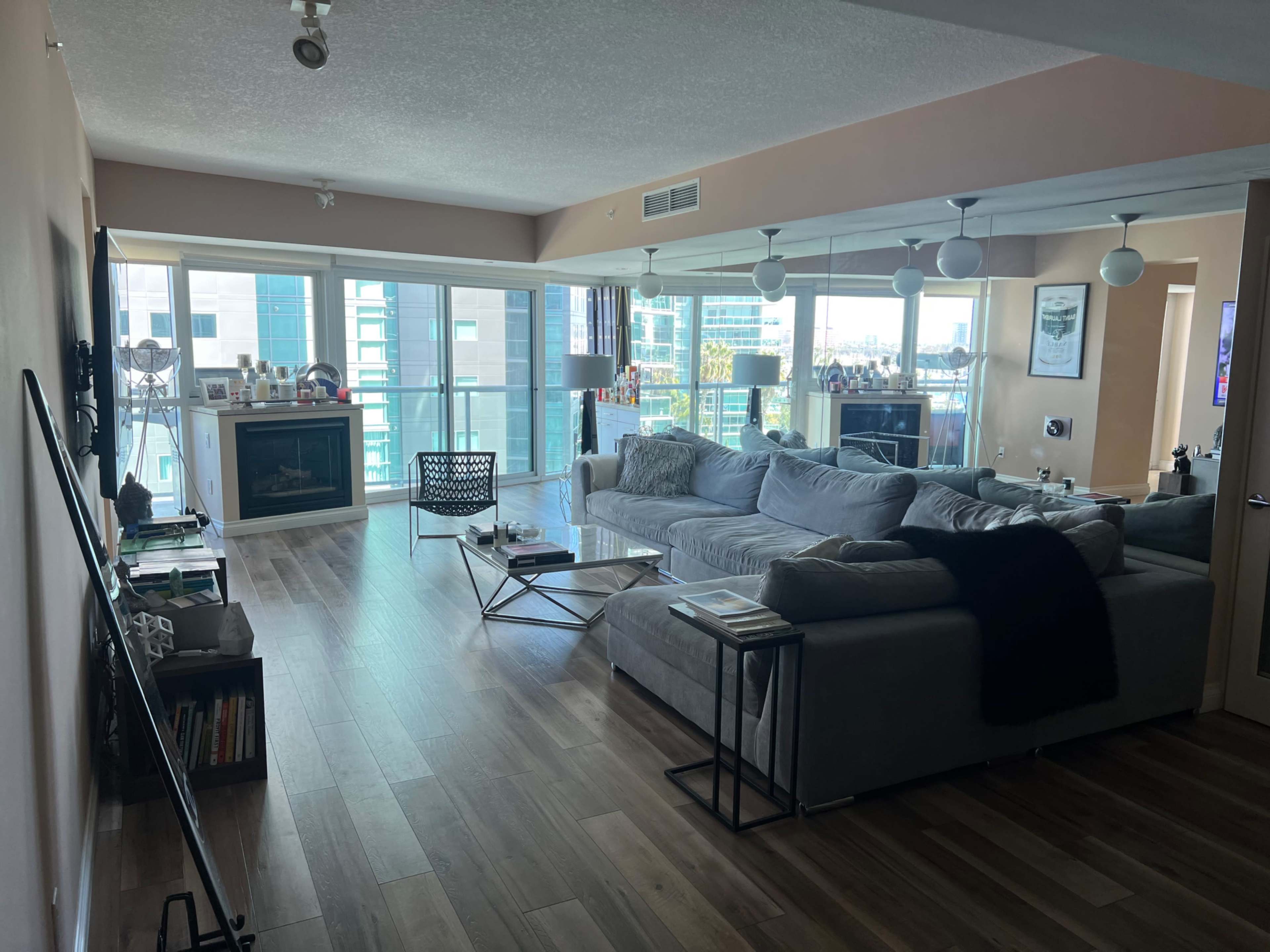 A modern living room with large windows, a sectional sofa, a glass coffee table, and a TV mounted on the wall.