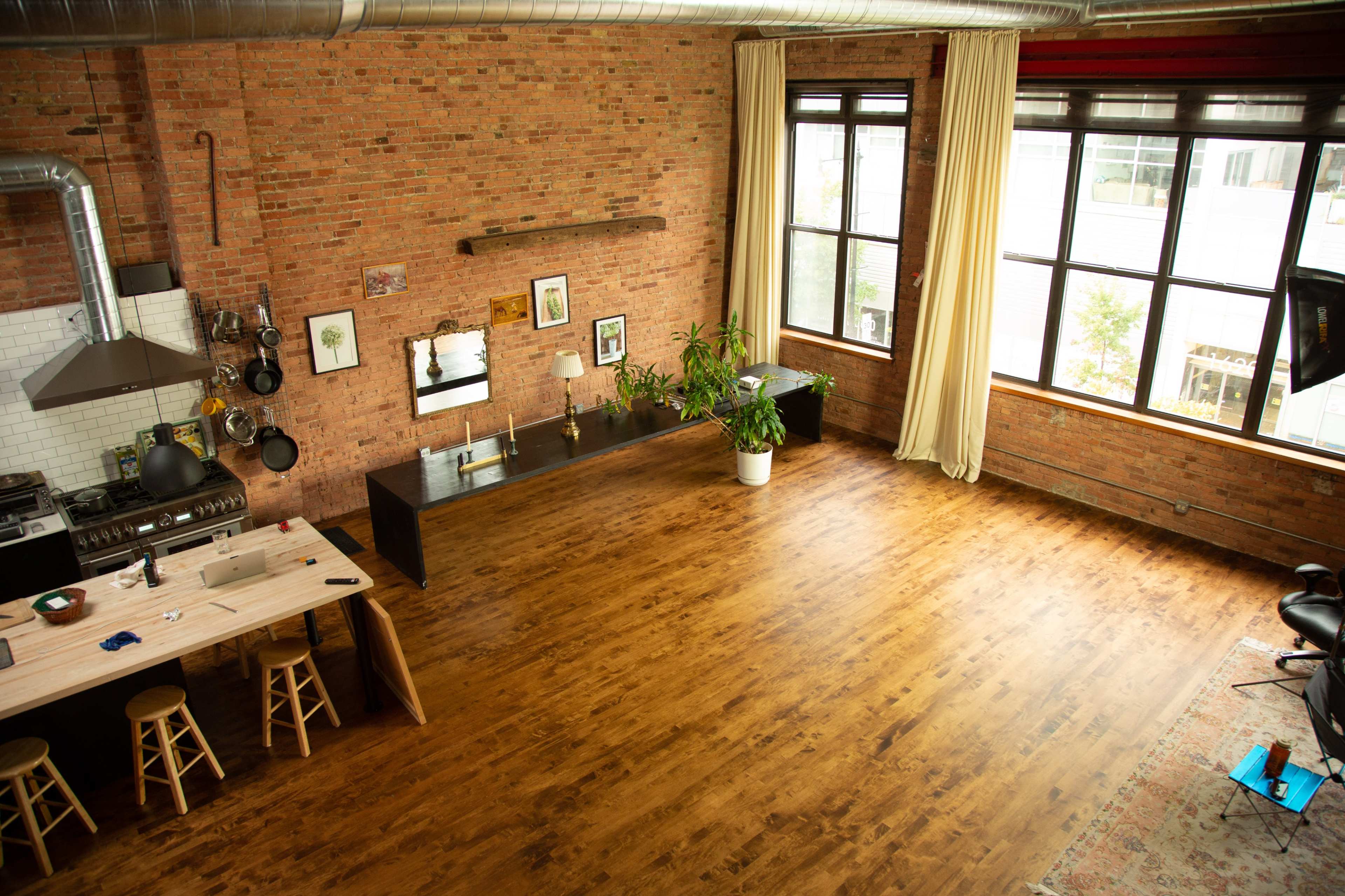 The image shows a spacious loft with exposed brick walls, a wooden floor, a kitchen area with a large stove and a wooden table, and large windows allowing natural light to enter.