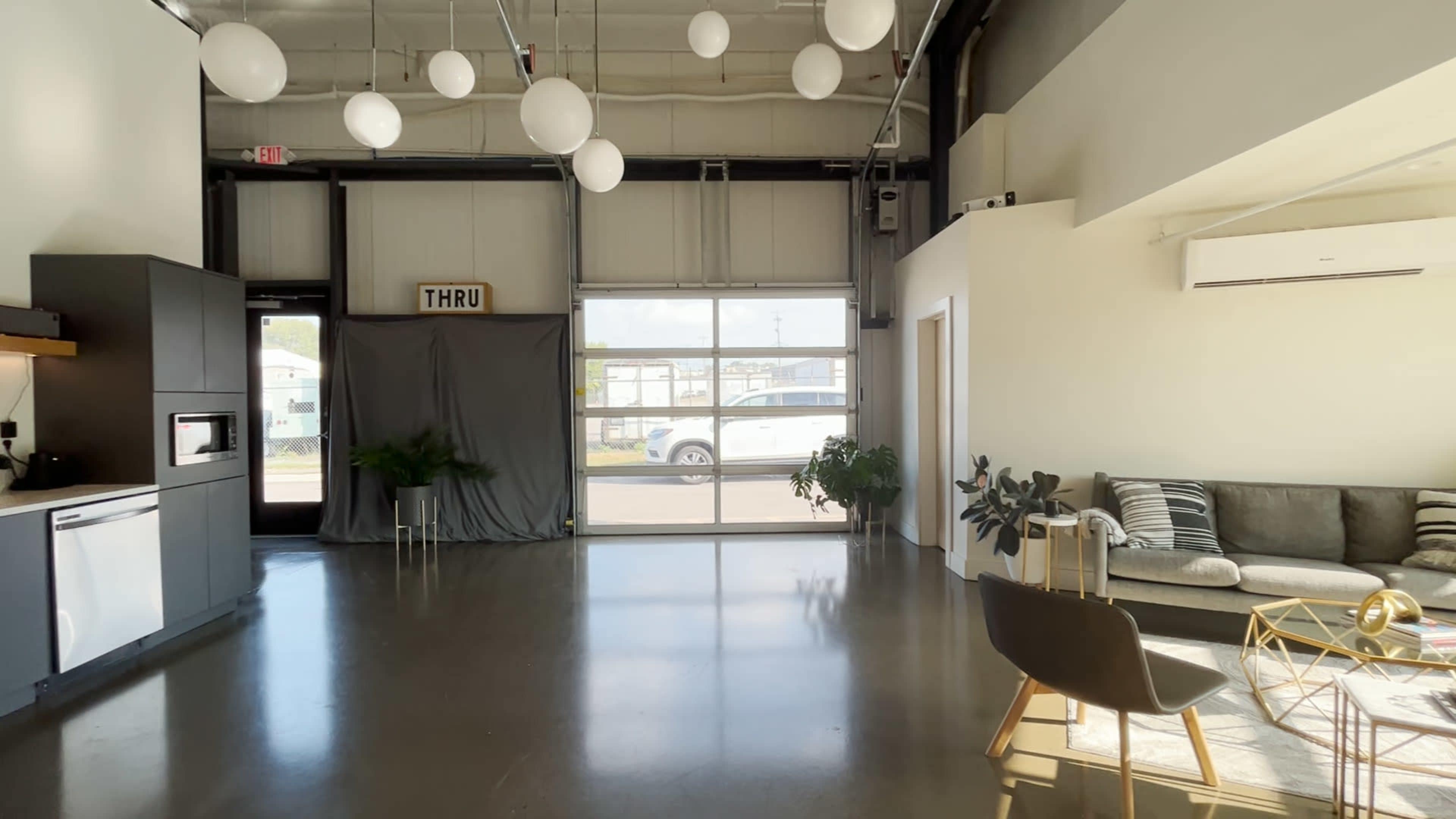 An interior space with modern decor, featuring a small kitchen area, a living area with a sofa, and large windows letting in natural light.