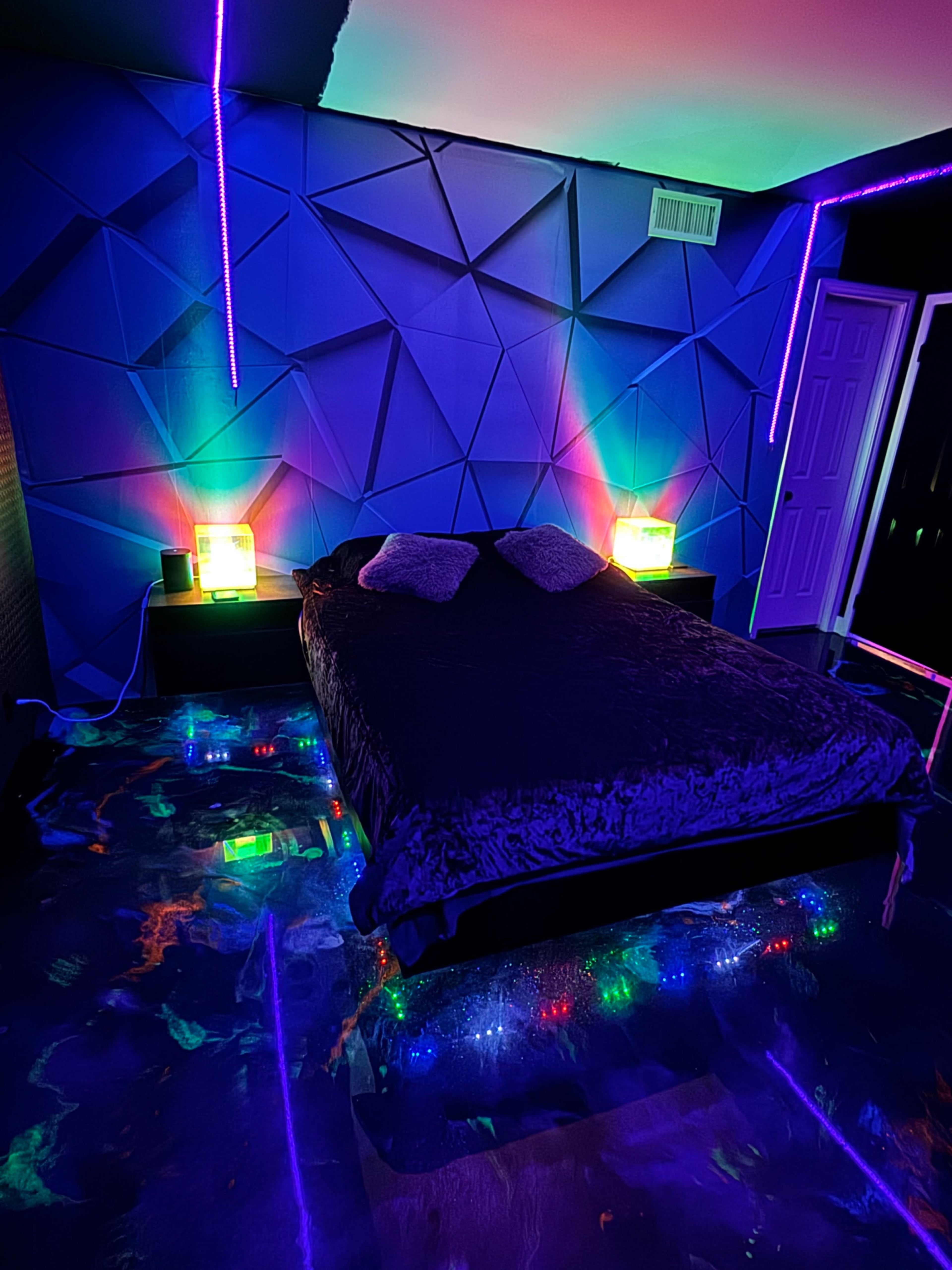 A bedroom with a geometric blue wall design, a dark bedspread on the bed, and colorful lights illuminating the space.