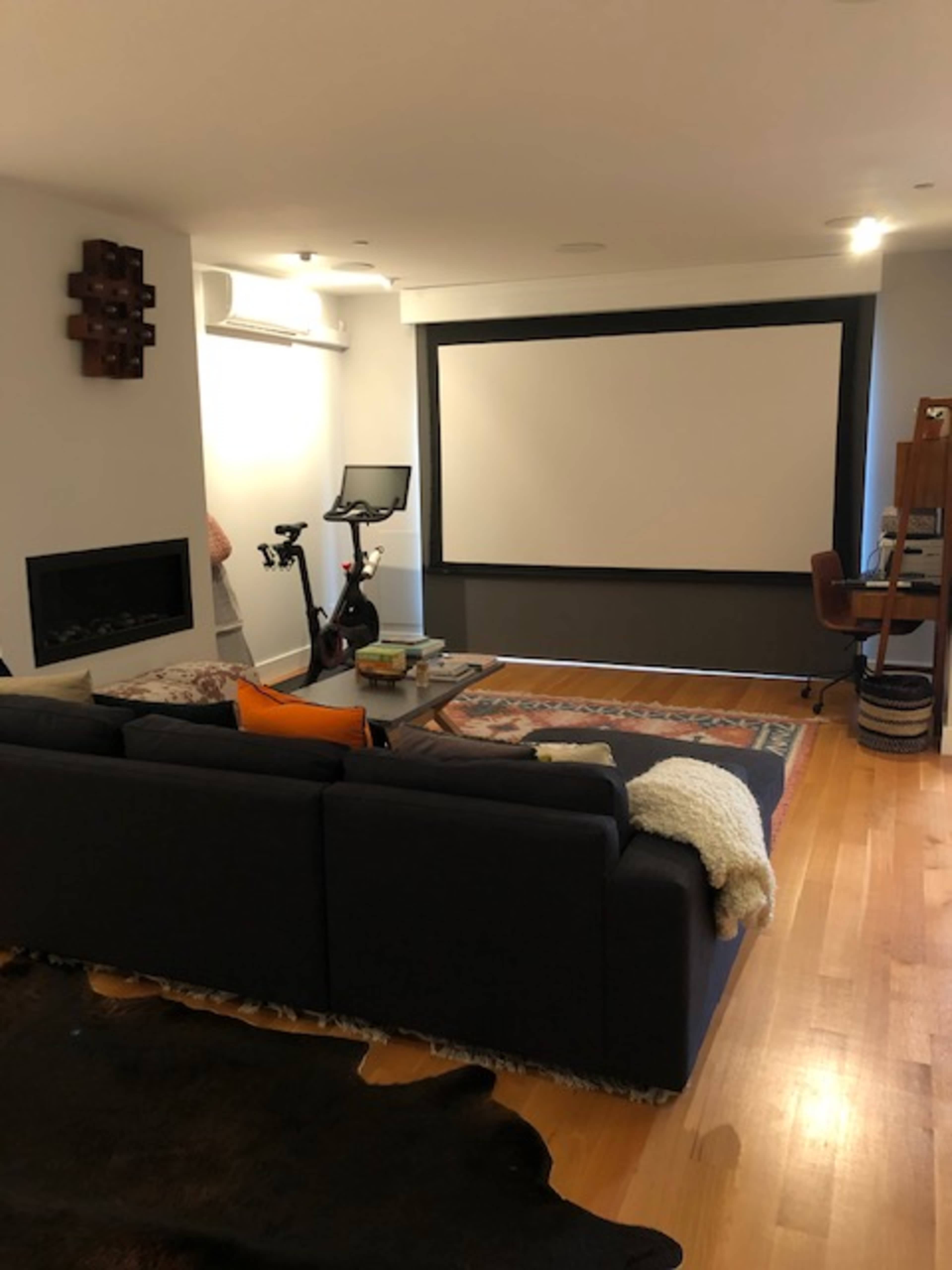 The image shows a living room with a sectional couch, a large screen or projector setup, a stationary bike, and a small desk.