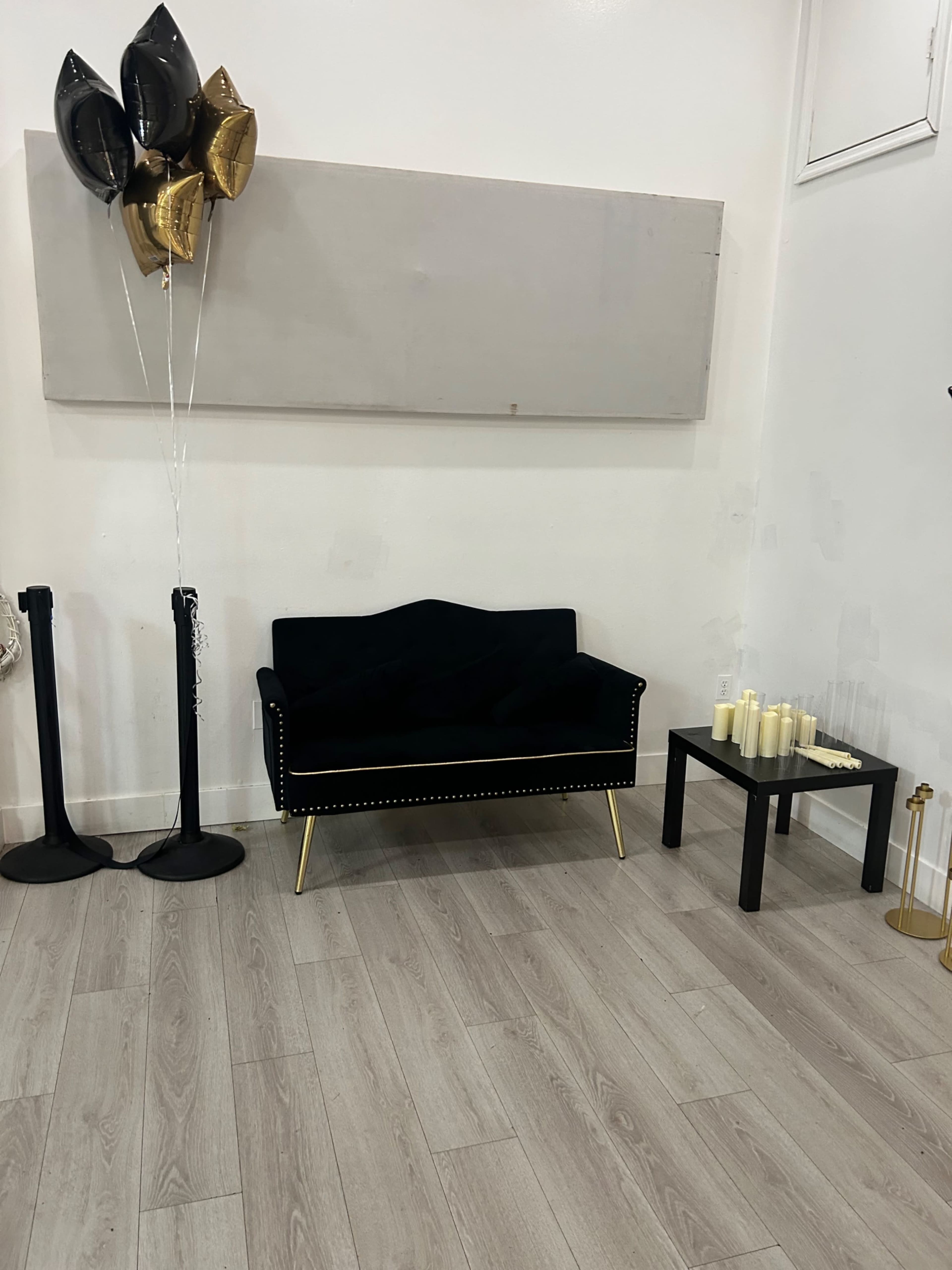 The image shows a black velvet loveseat with gold accents beside a small black table, candles, and black and gold balloons in a simple interior space.
