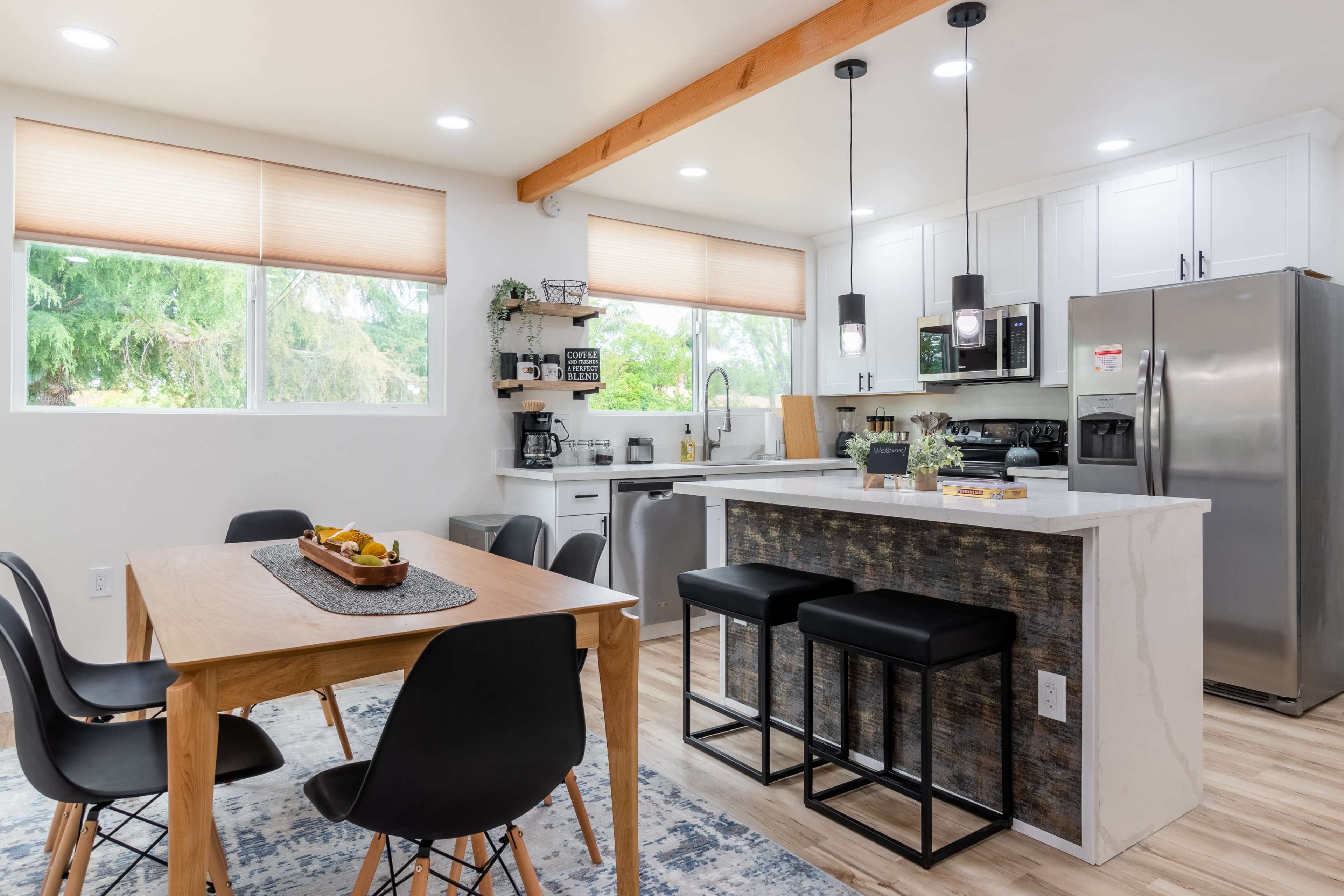 The image shows a modern kitchen and dining area featuring a wooden table with black chairs, a countertop with bar stools, and large windows allowing natural light to enter.