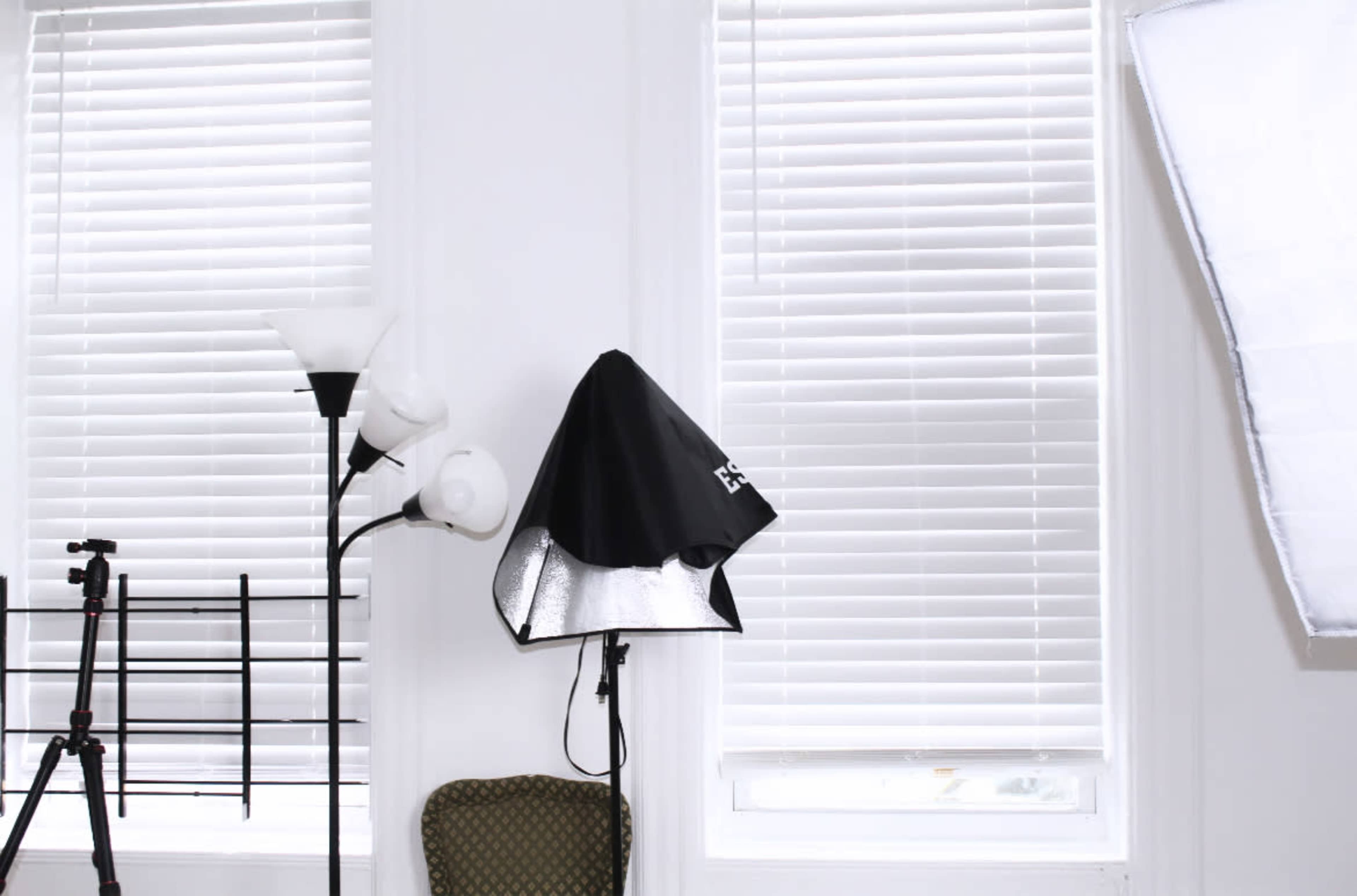A lighting setup is positioned near a window with closed blinds, along with a tripod and a lamp in the room.