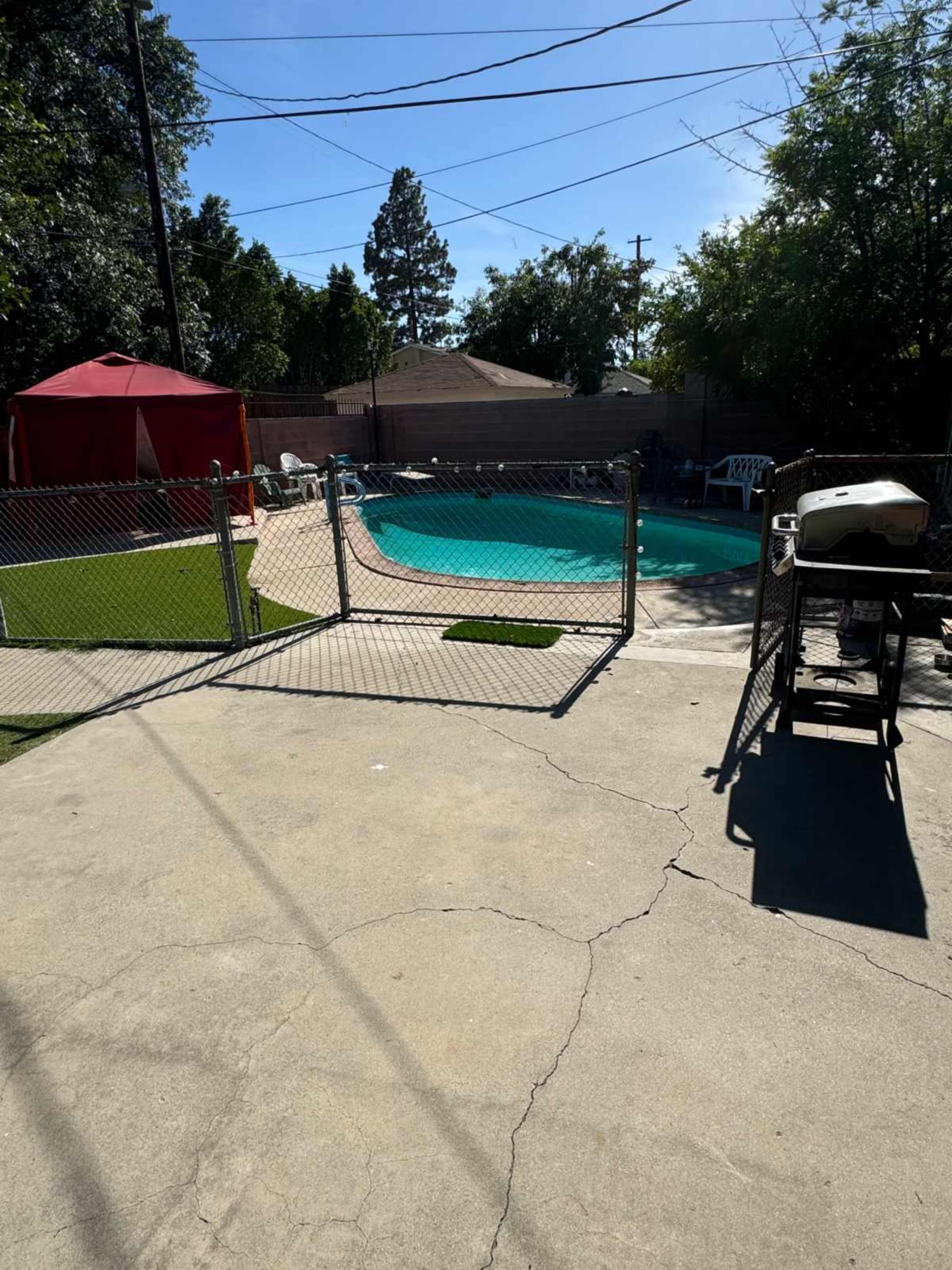 The image shows a swimming pool surrounded by a fence, with a red canopy tent and seating area nearby, under a clear blue sky.
