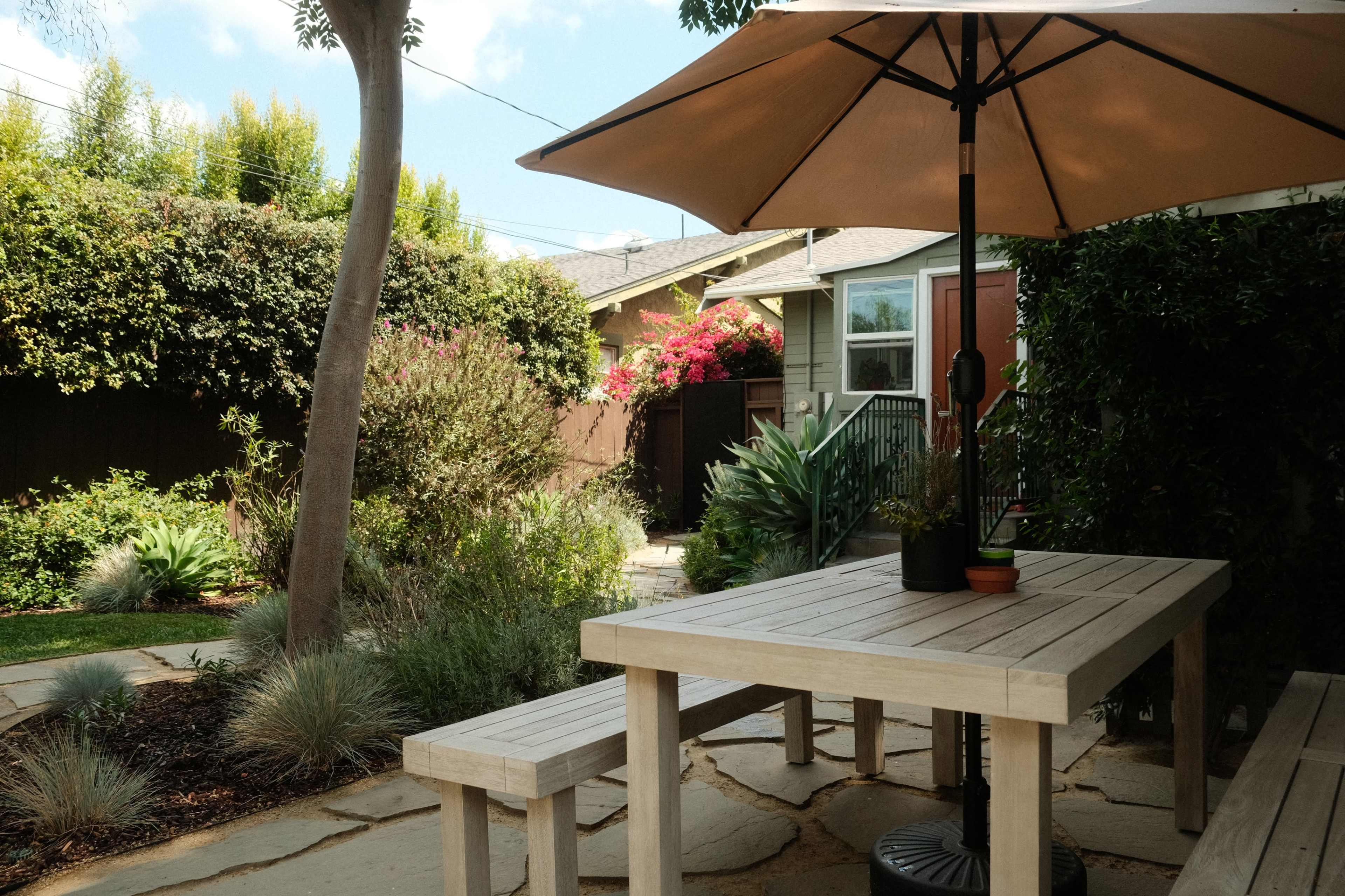 A wooden table and bench are set up under a large umbrella in a landscaped backyard surrounded by greenery and flowering plants.