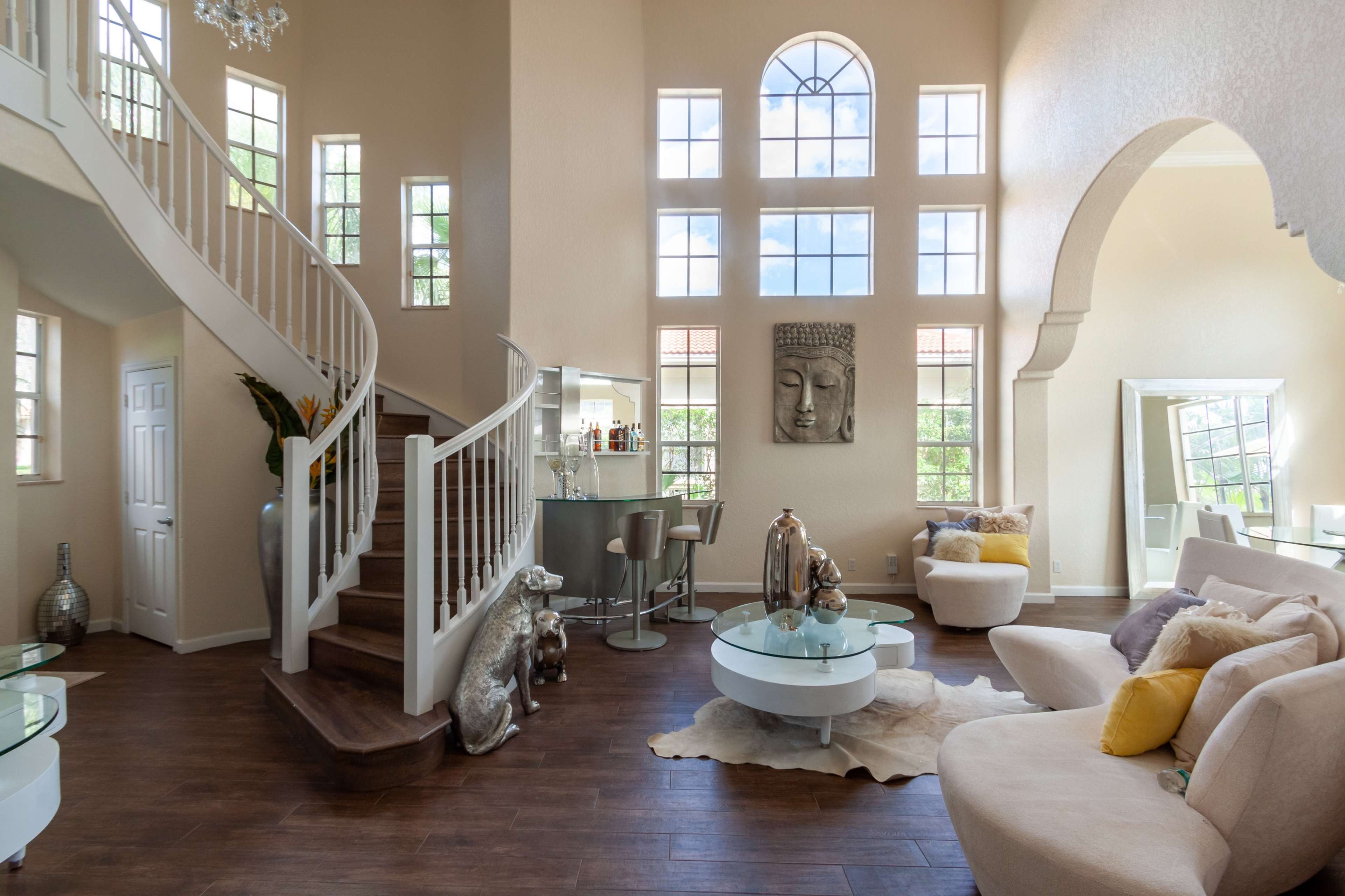 A spacious living room features large windows, a curved staircase, and modern furniture arranged around a coffee table.