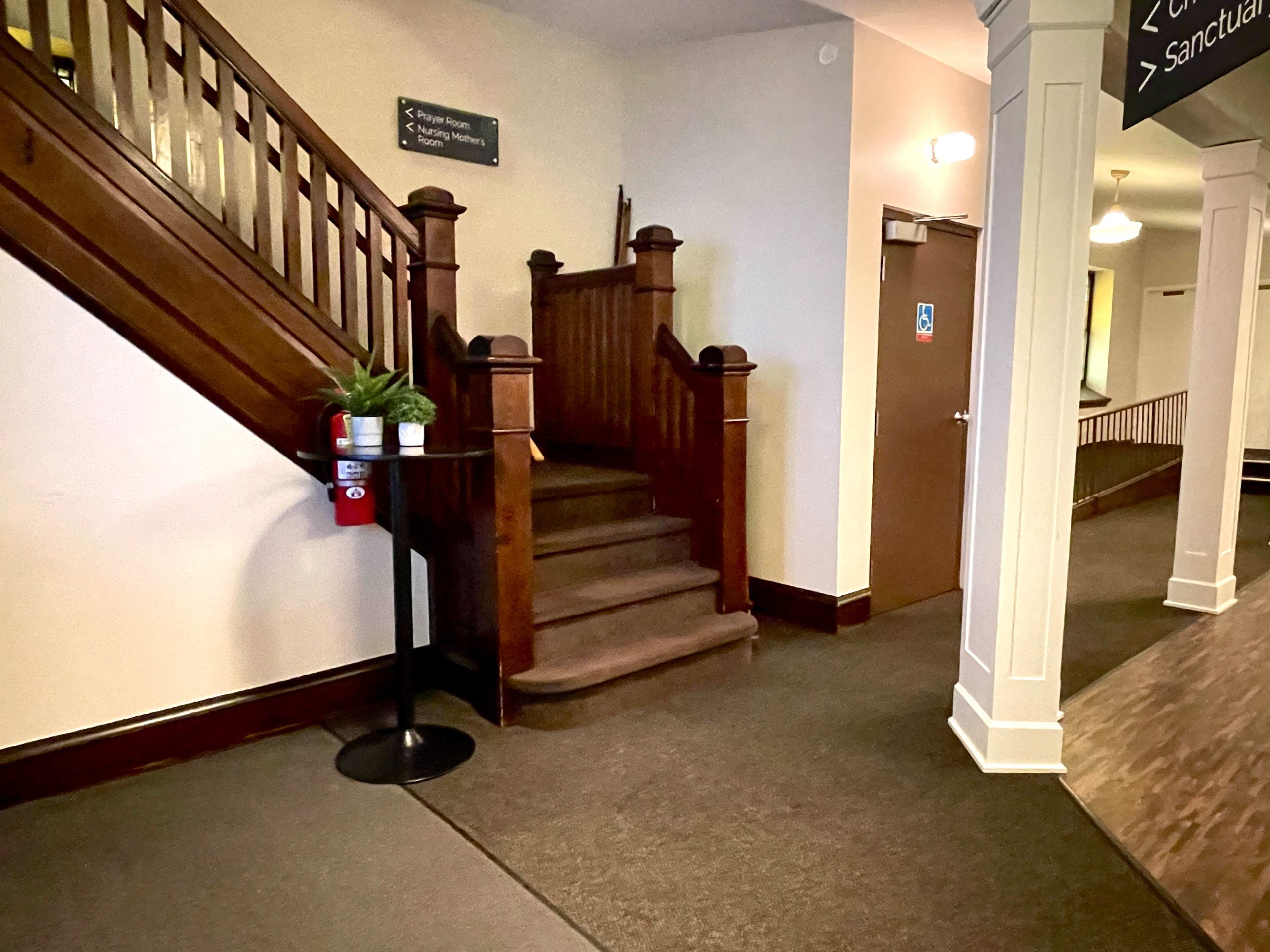 A wooden staircase at the end of a hallway with a plant on a stand beside it and signs indicating directions.