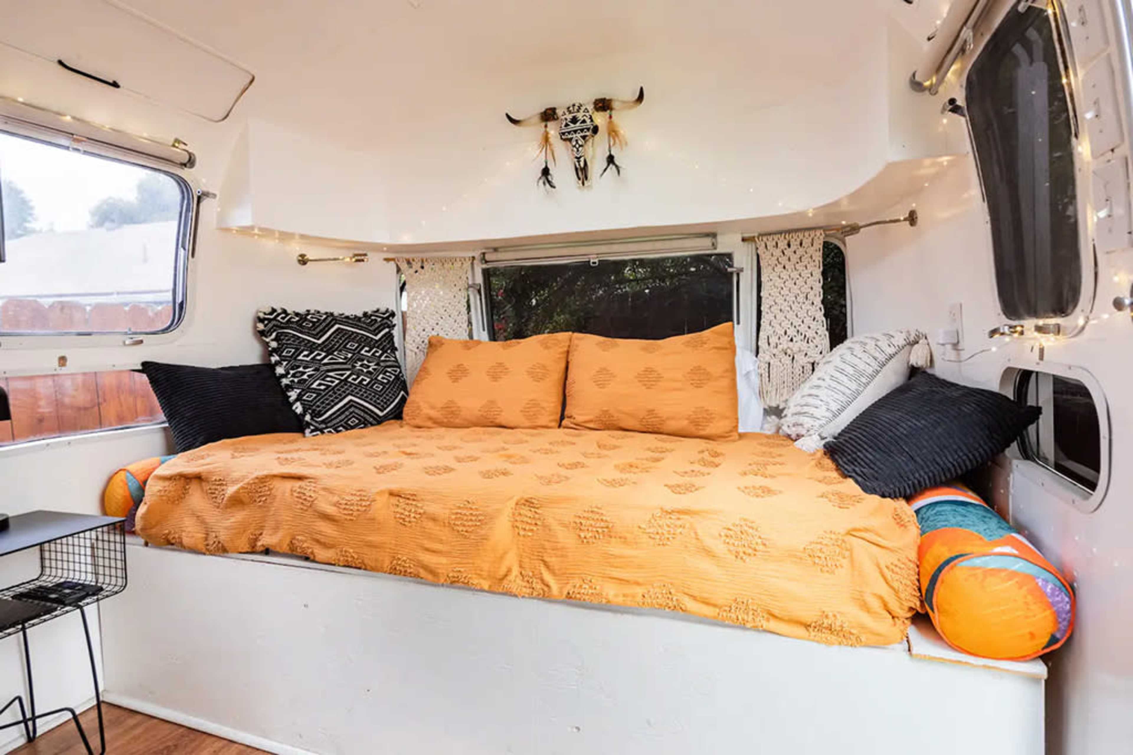The image shows a cozy interior of a small trailer featuring a bed with an orange blanket, several pillows, and decorative elements like string lights and a patterned wall hanging.