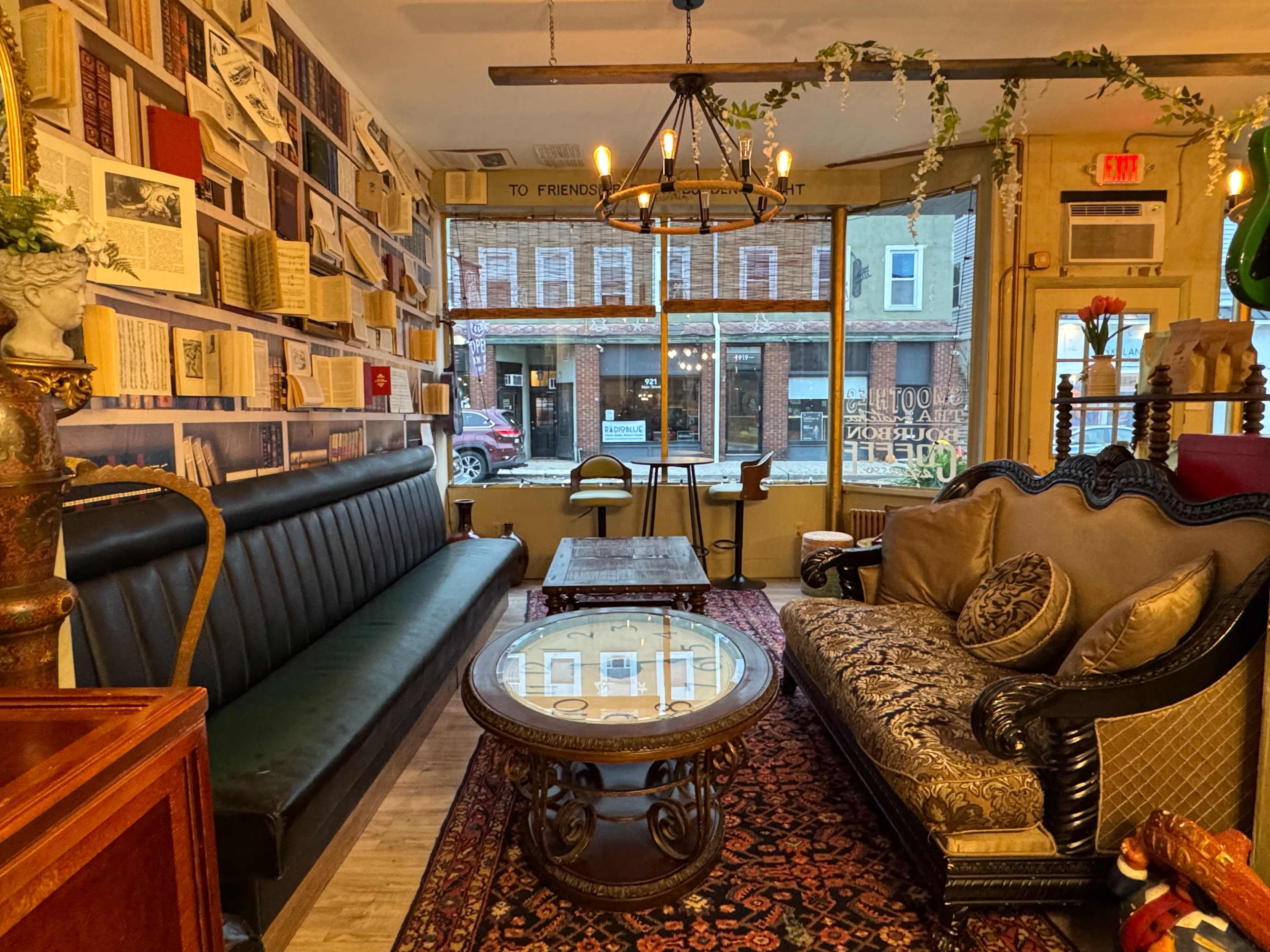 The image shows a cozy café interior with two different seating areas, one featuring a black leather sofa and the other a decorative upholstered couch, surrounded by bookshelves and large windows.