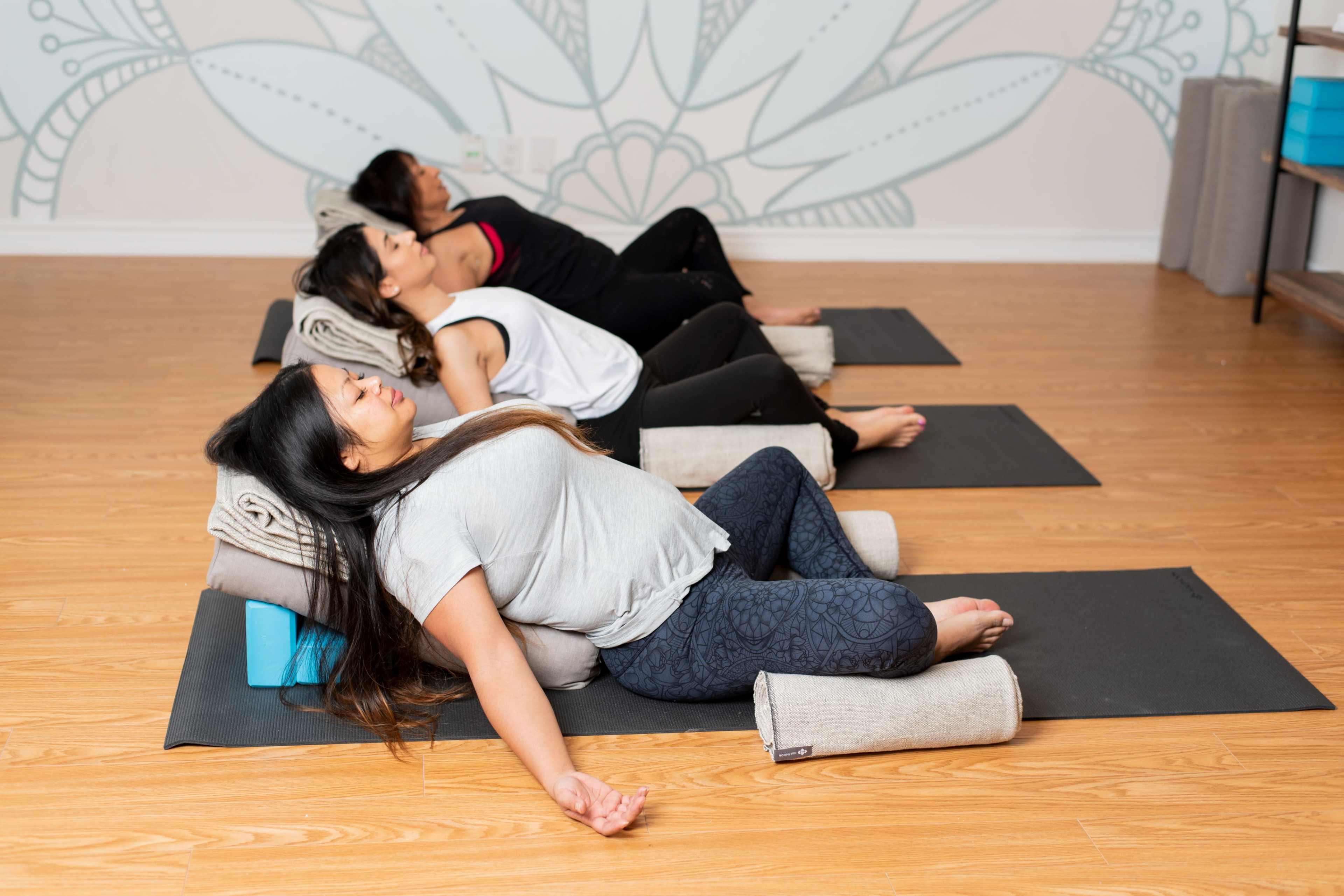 Three women practice restorative yoga on mats in a studio with a decorative wall mural.