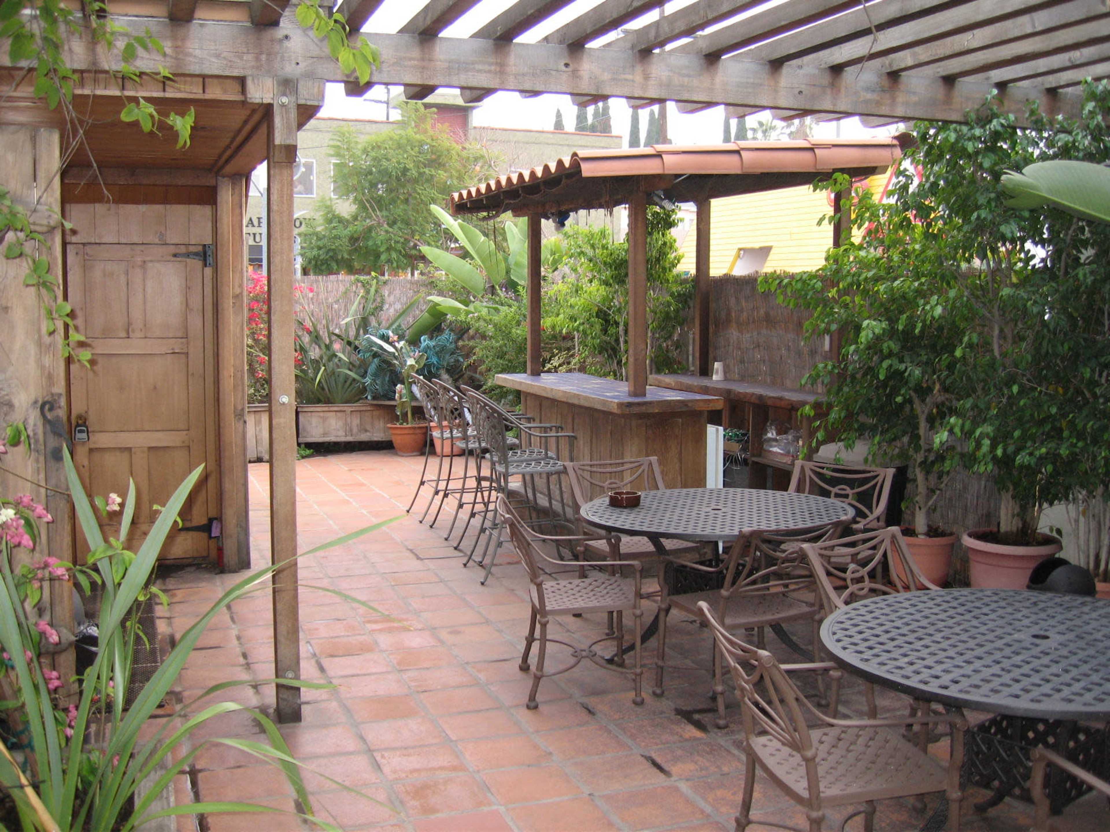 The image shows a spacious patio with several metal tables and chairs, surrounded by greenery and featuring a wooden bar under a pergola.